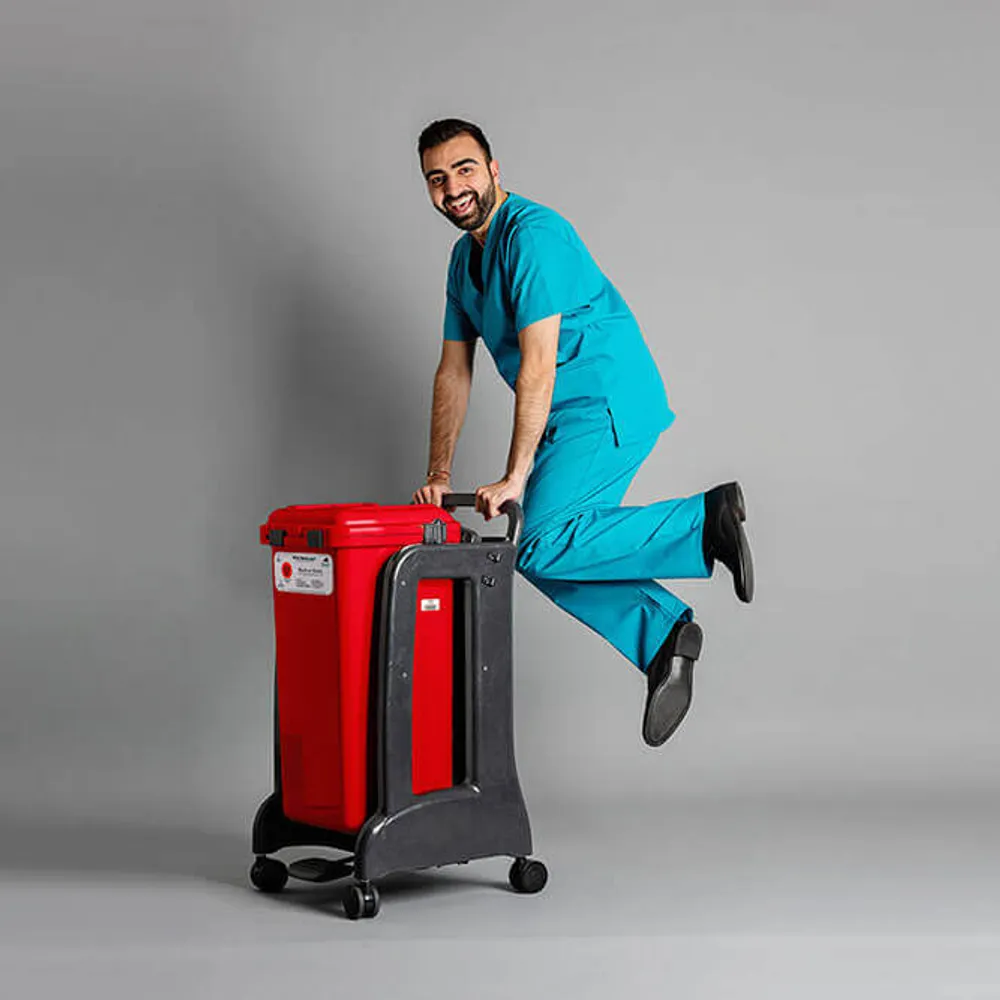 Hospital staff member in teal scrubs playfully riding a Daniels Health's red clinical waste bin on wheels in studio