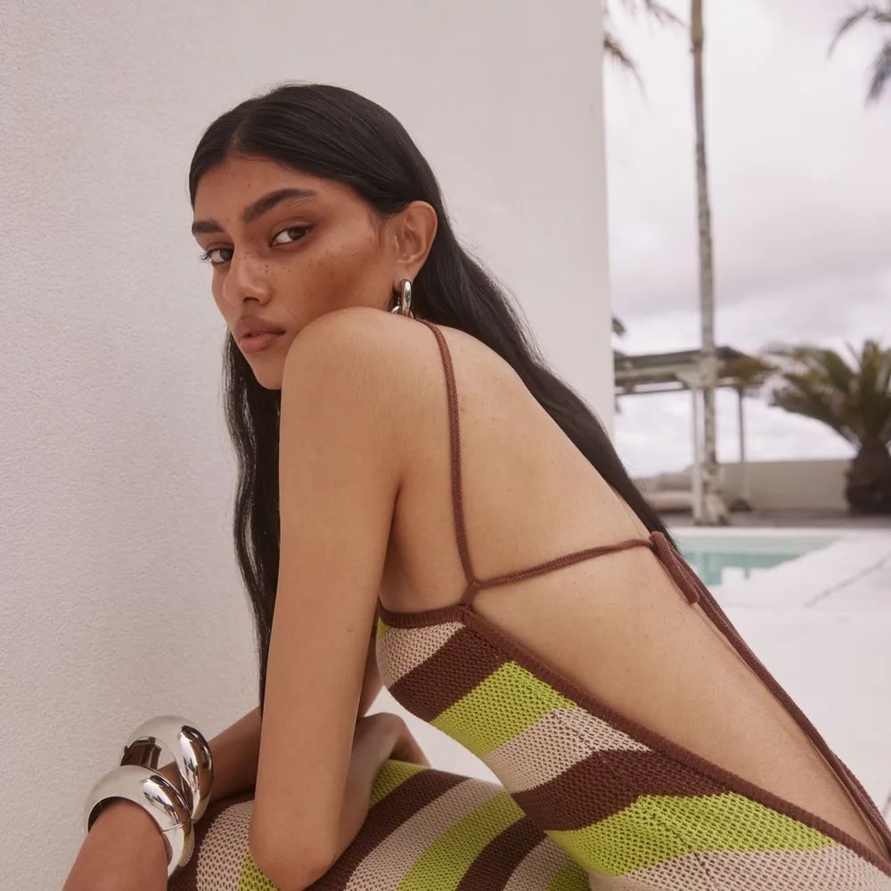 Model in Runaway The Label striped knit dress with low back and thin straps in brown, lime and beige tones styled with bold silver cuff bracelet by the poolside.