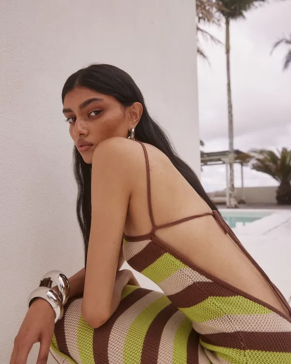 Model in Runaway The Label striped knit maxi dress with low open back and thin straps in brown, lime and beige tones styled poolside with silver cuff bracelet