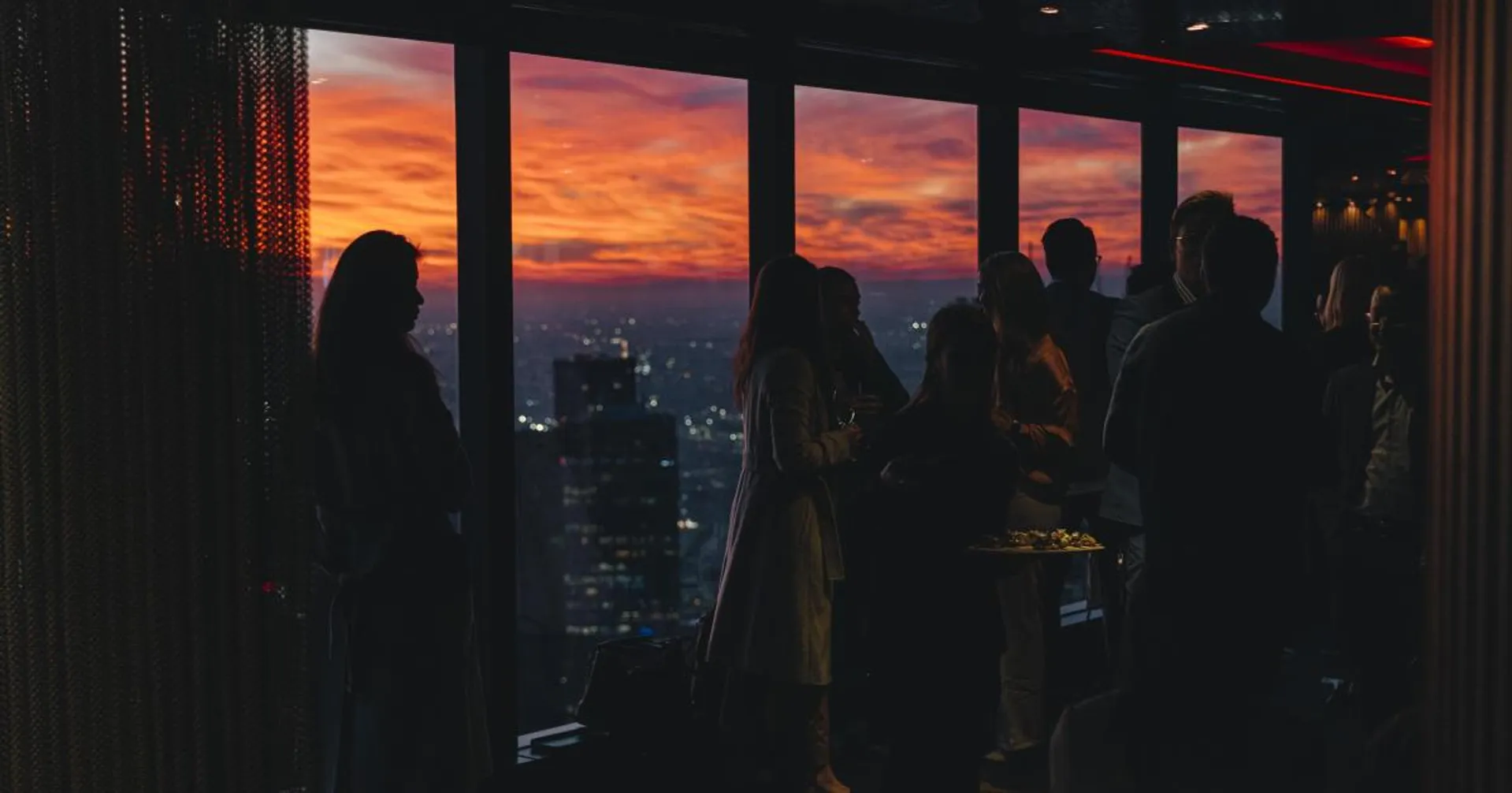 Silhouettes of professionals networking at a healthcare event, set against a vibrant city sunset through floor-to-ceiling windows.