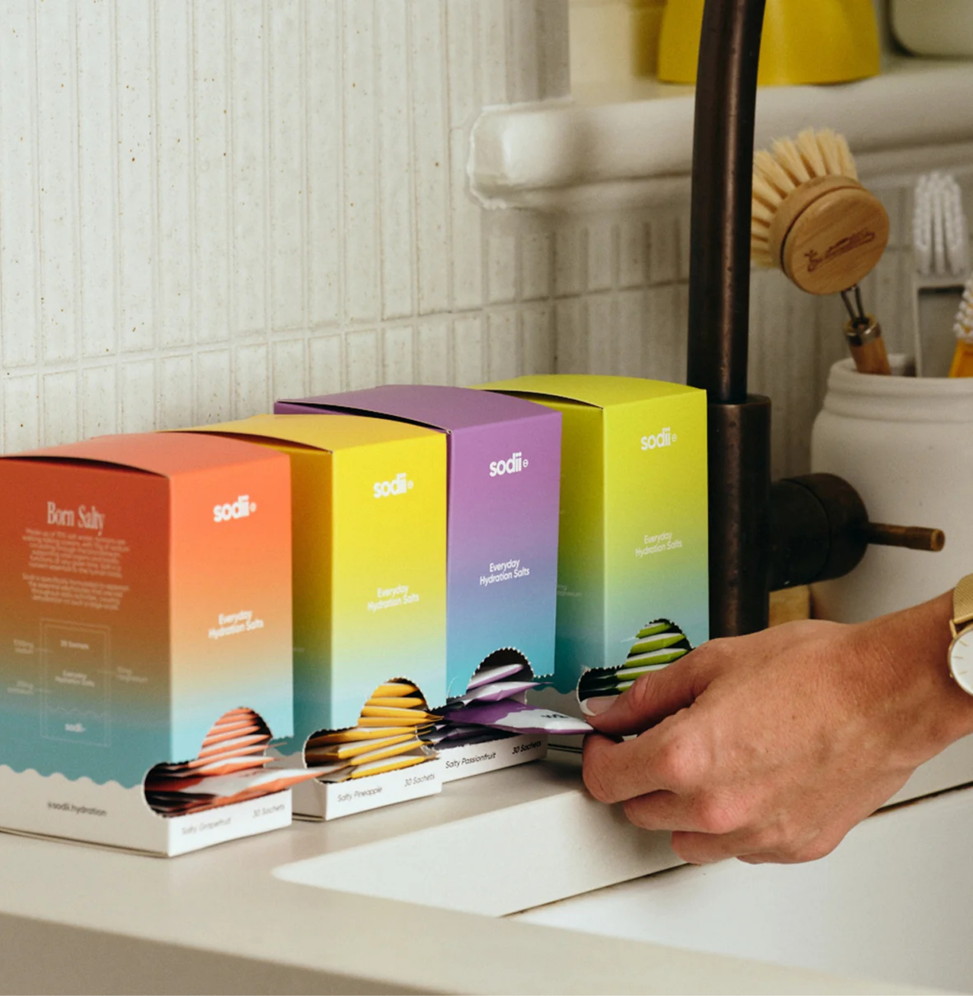 Hand reaching for a Sodii hydration sachet from a colourful kitchen bench display of Sodii electrolyte boxes in gradient packaging.