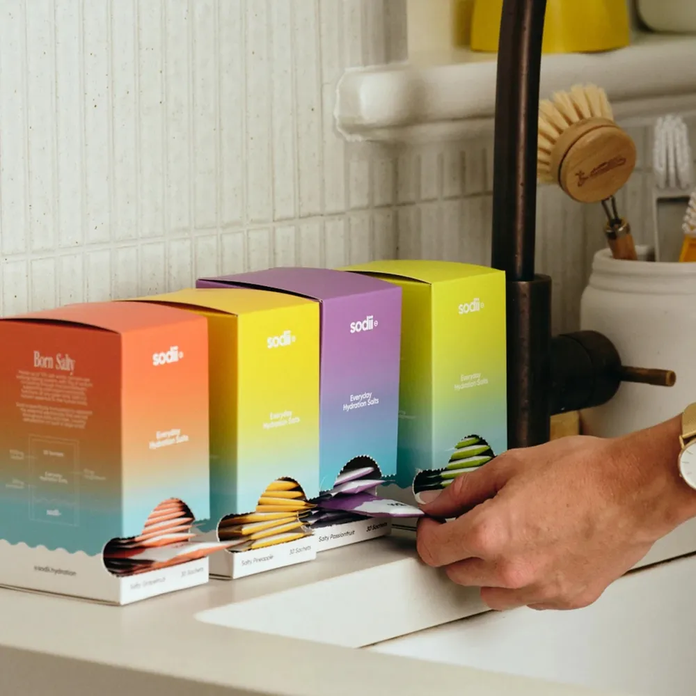 Hand reaching for a Sodii hydration sachet from a colourful kitchen bench display of Sodii electrolyte boxes in gradient packaging.