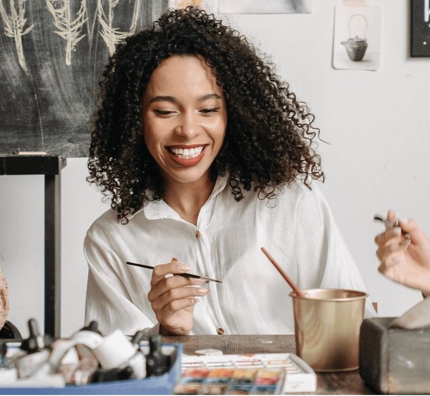 Smiling woman painting during a WeTeachMe creative class, holding a brush at a studio table. 