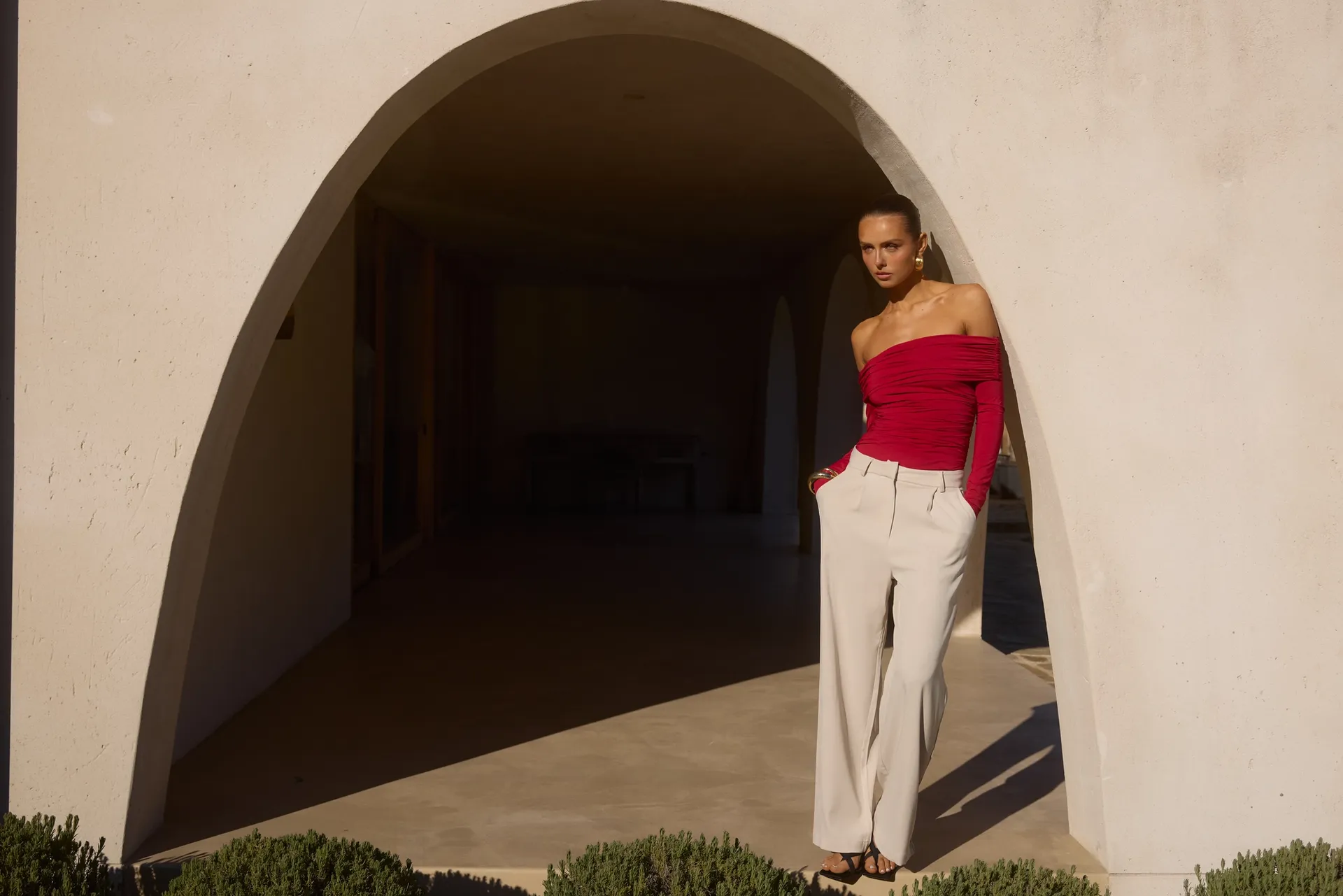 Model wearing Runaway The Label off-shoulder red ruched top with wide-leg cream trousers styled under modern arched architecture