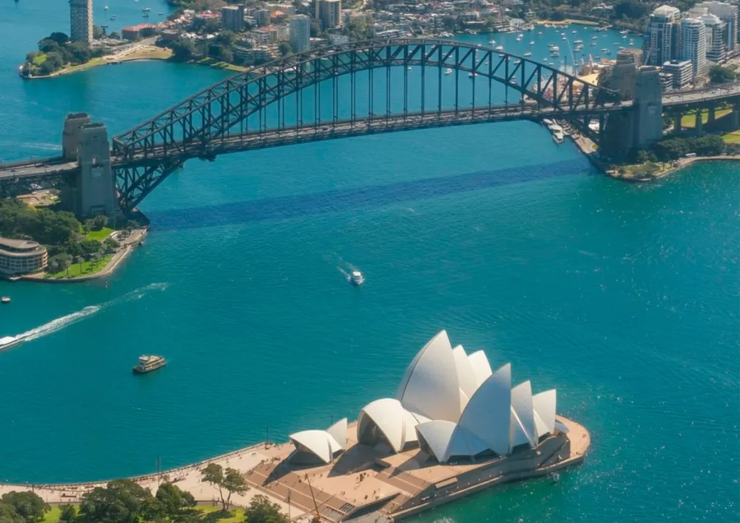 Aerial view of Sydney Harbour showing the Sydney Opera House in the foreground and the Harbour Bridge arching across the sparkling blue water