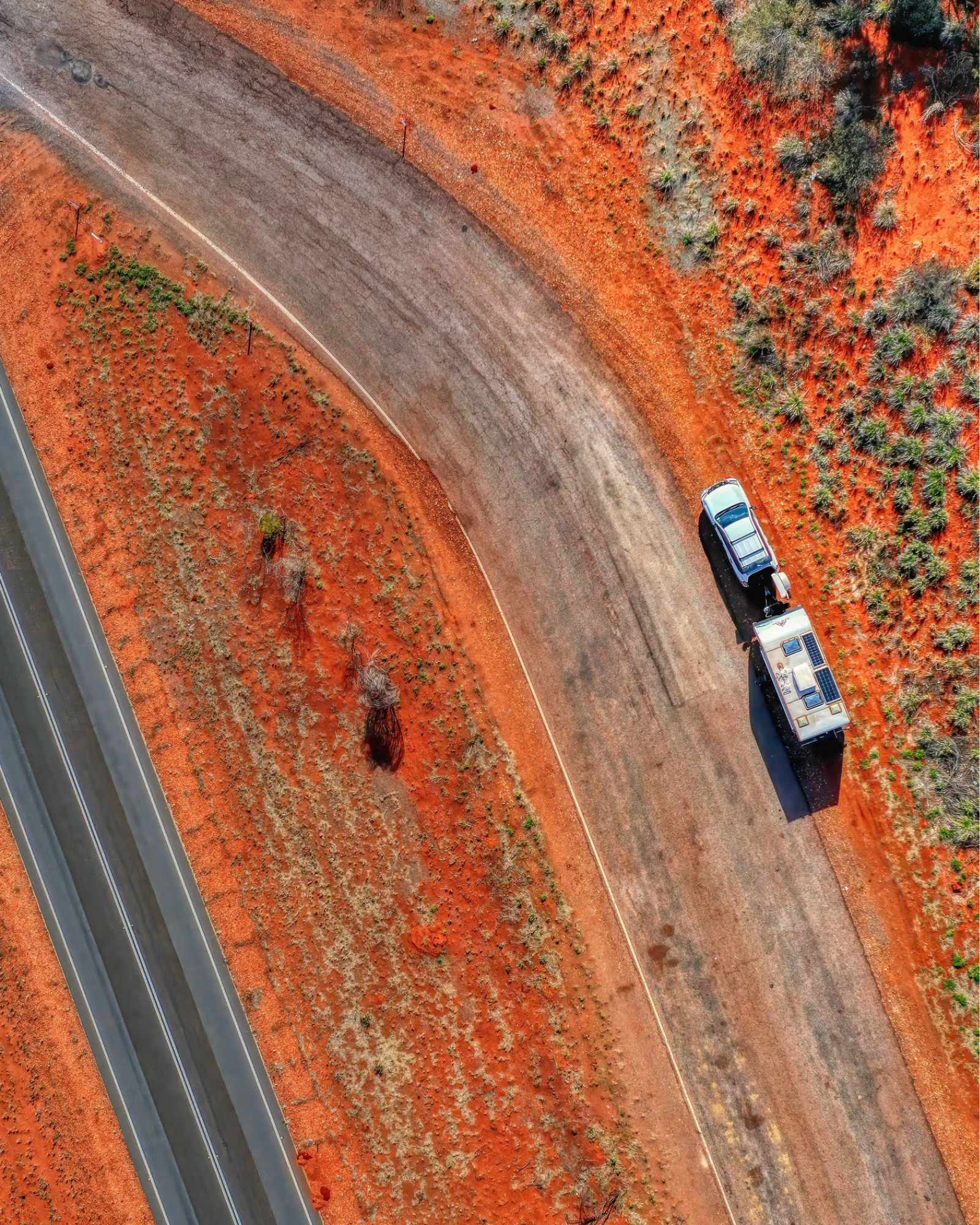 Club 4x4 vehicle towing a caravan along a winding red-earth outback road captured from above.