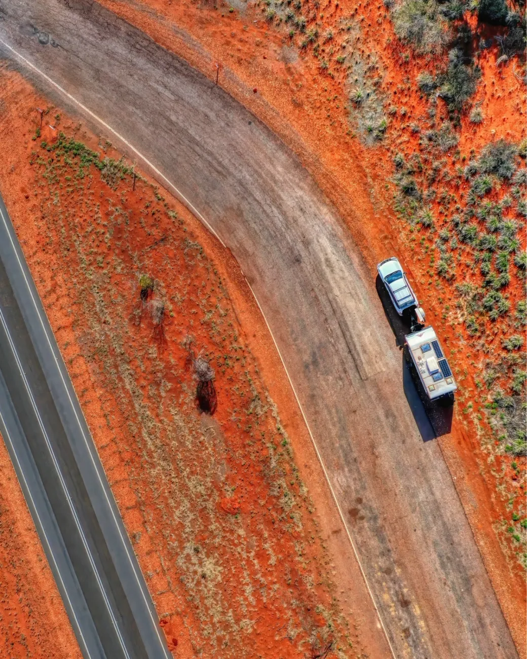 Club 4x4 vehicle towing a caravan along a winding red-earth outback road captured from above.