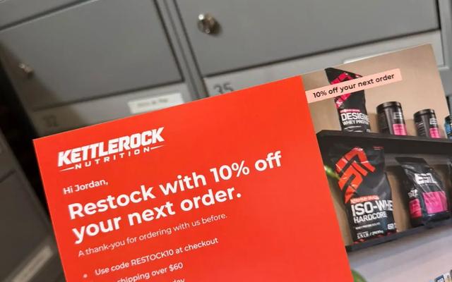 Hand holding a red reactivation postcard offering 10% off a returning customer's next order, with a QR code to reorder, in front of mailboxes