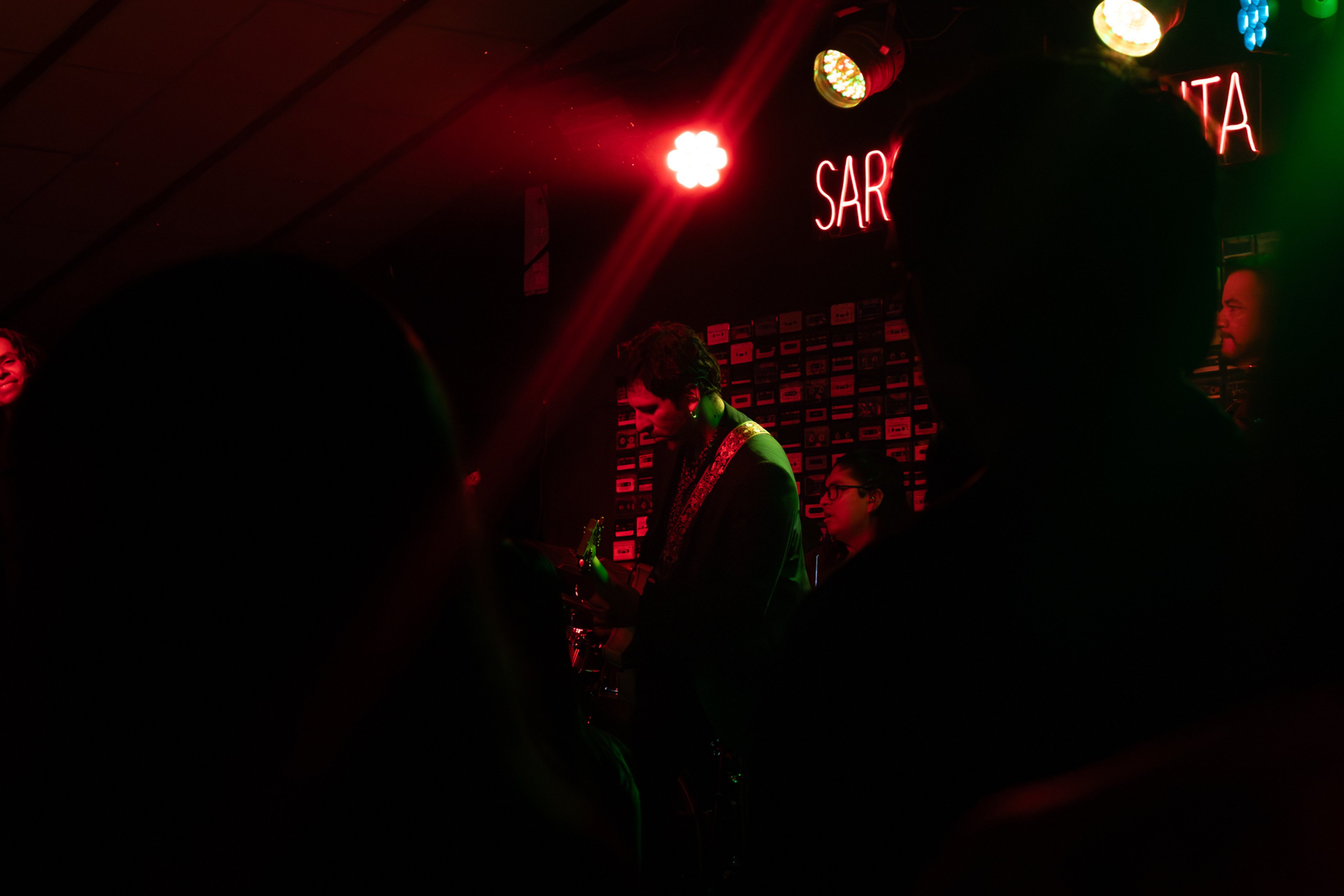 Banda tocando en un escenario pequeño con luces rojas y verdes, vista desde entre el público.