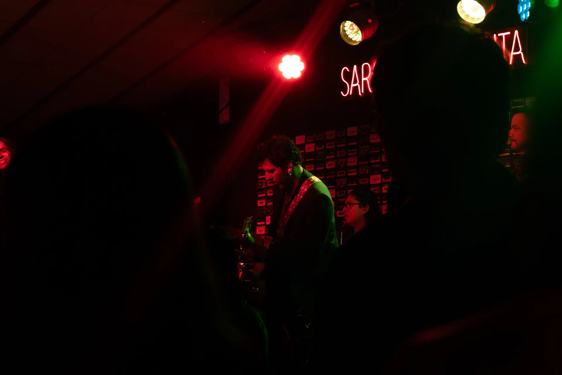 Banda tocando en un escenario pequeño con luces rojas y verdes, vista desde entre el público.