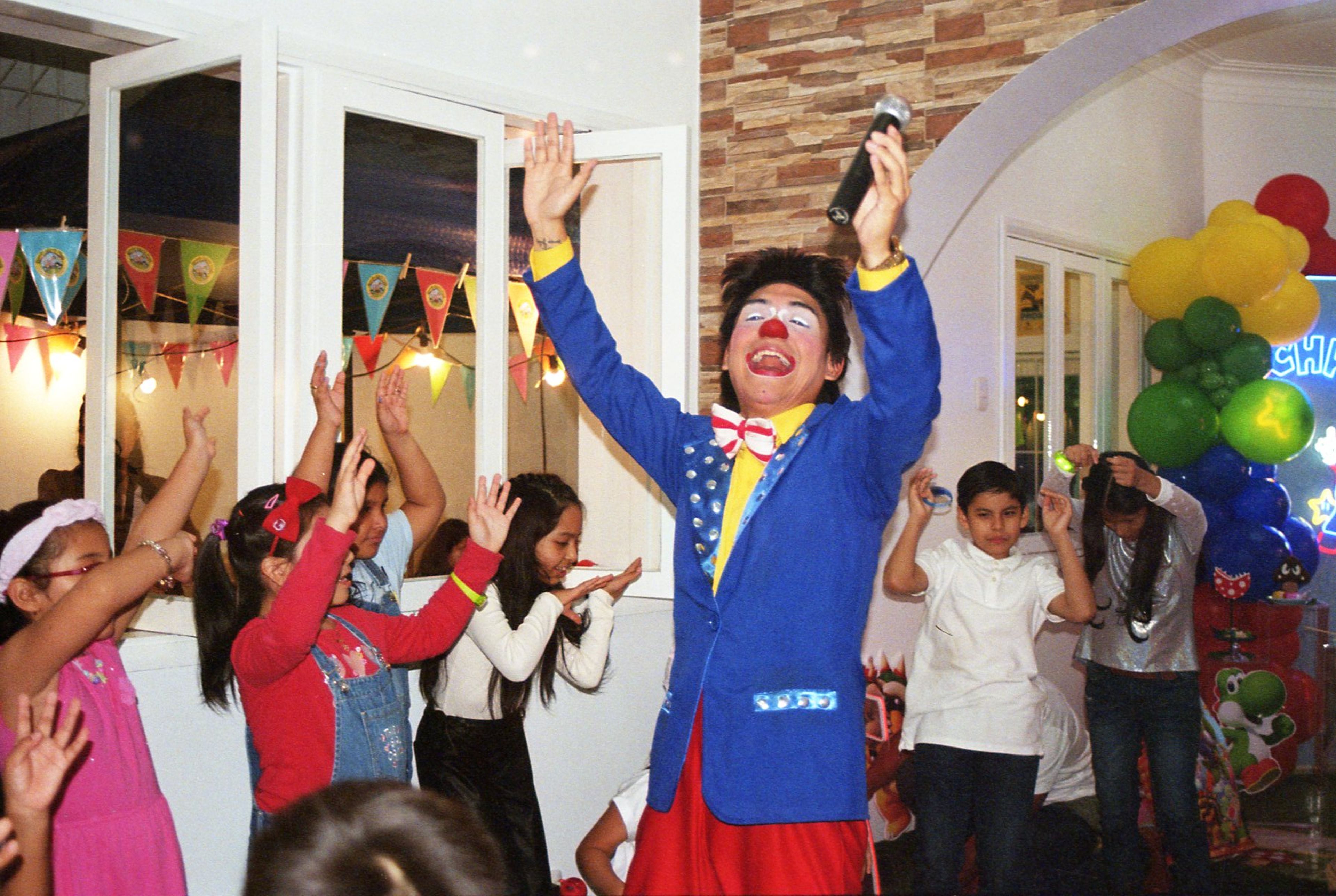 Animador disfrazado de payaso dirigiendo un juego con varios niños levantando los brazos en una fiesta