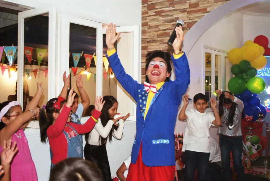 Animador disfrazado de payaso dirigiendo un juego con varios niños levantando los brazos en una fiesta