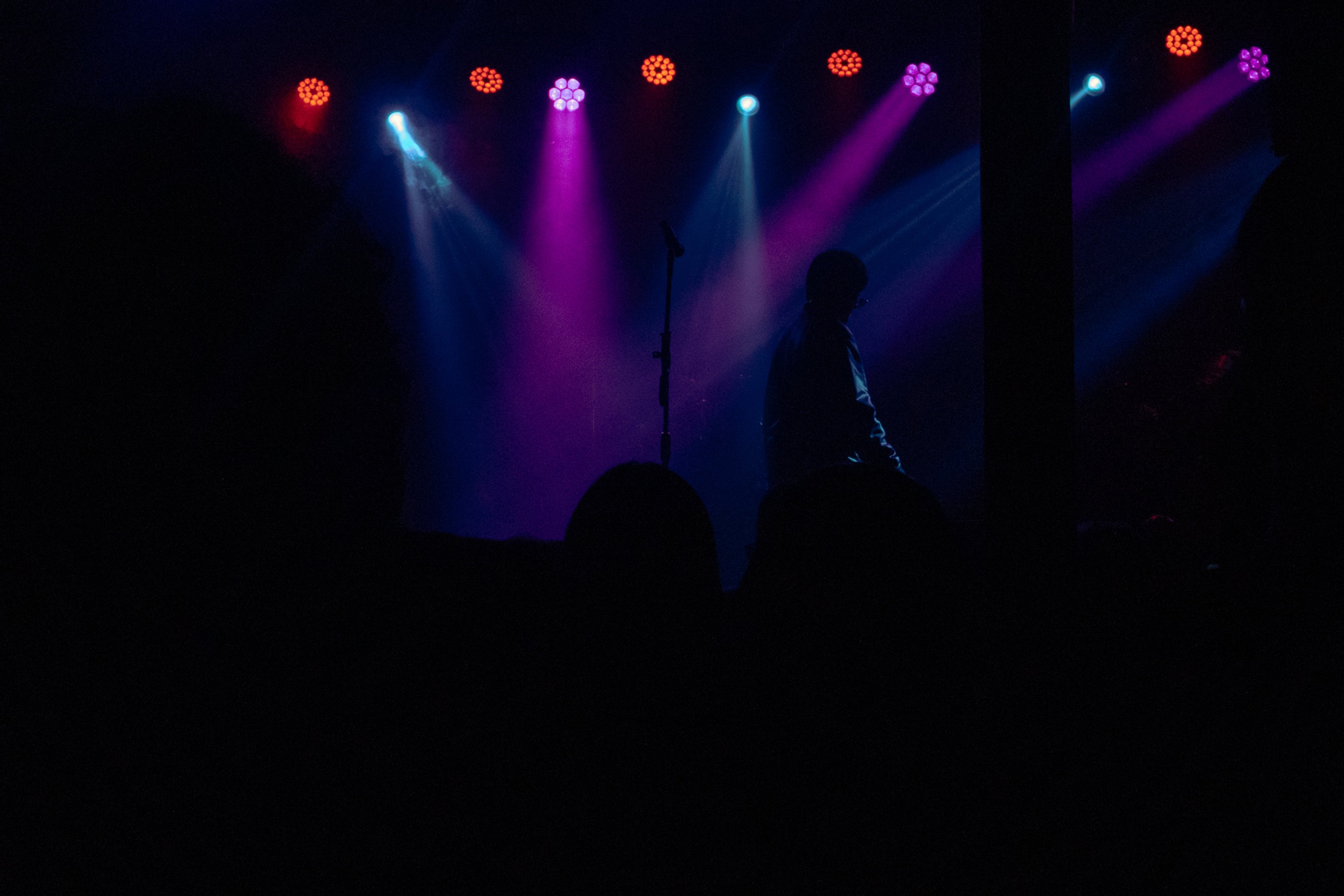 Silueta de un artista en el escenario bajo luces moradas y azules, con un micrófono al centro.