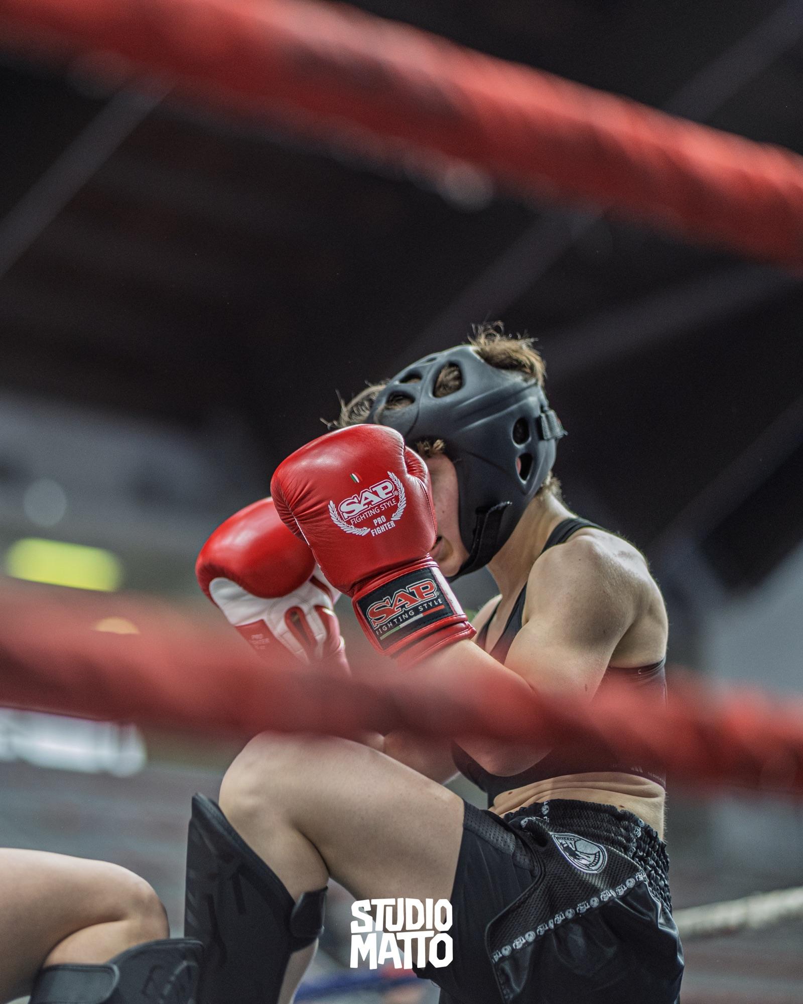 Una combattente si protegge il volto con i guantoni rossi, ripresa tra le corde del ring