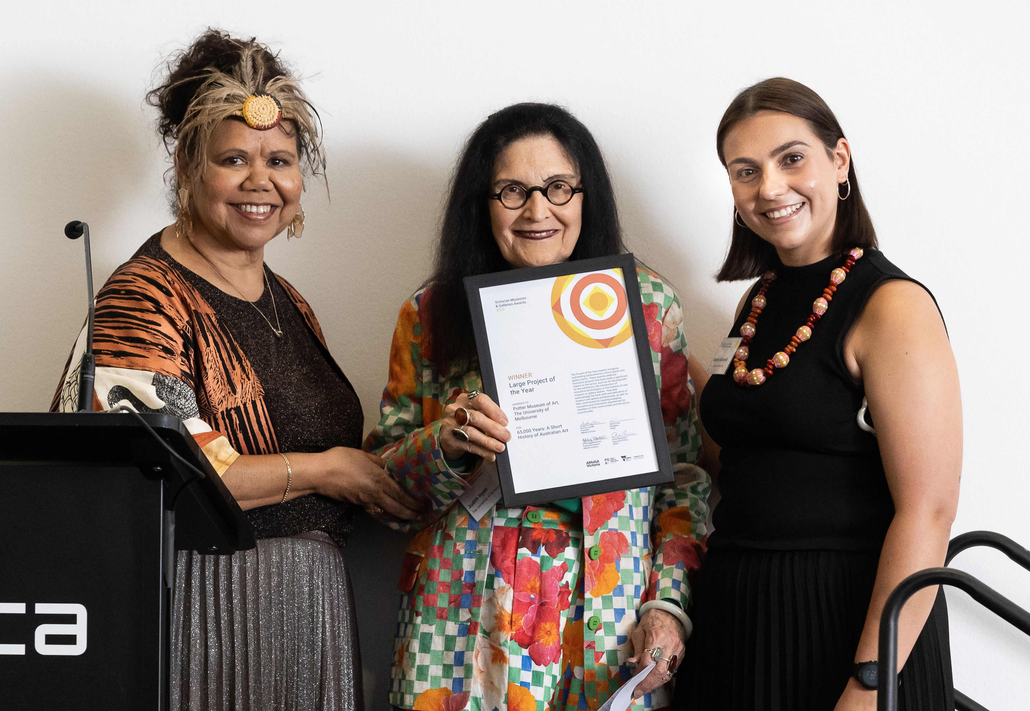 Three people stand together smiling; the person in the centre holds a framed certificate. They are dressed in colourful and patterned outfits, and are posing in front of a white wall.