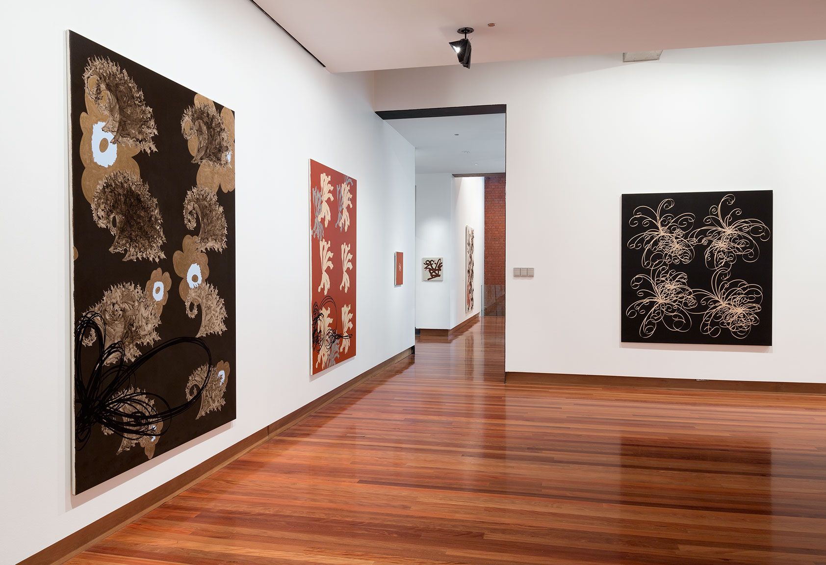 A photograph of a gallery space with three large abstract paintings in black, gold and dark red featuring repeated shapes. 