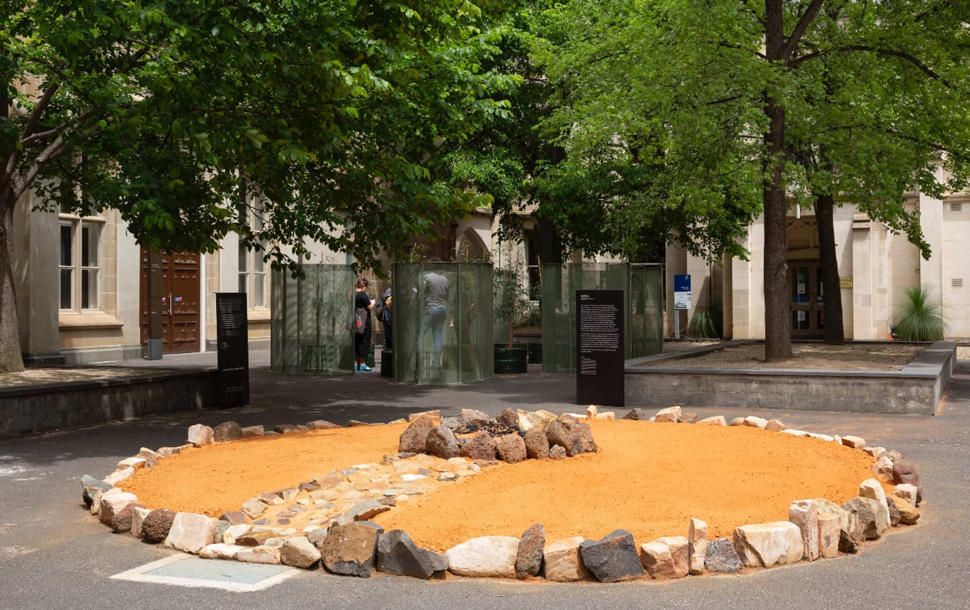 A circular outdoor installation with orange sand and rocks is arranged on the ground, surrounded by trees and informational black signs, in a courtyard with stone buildings in the background.