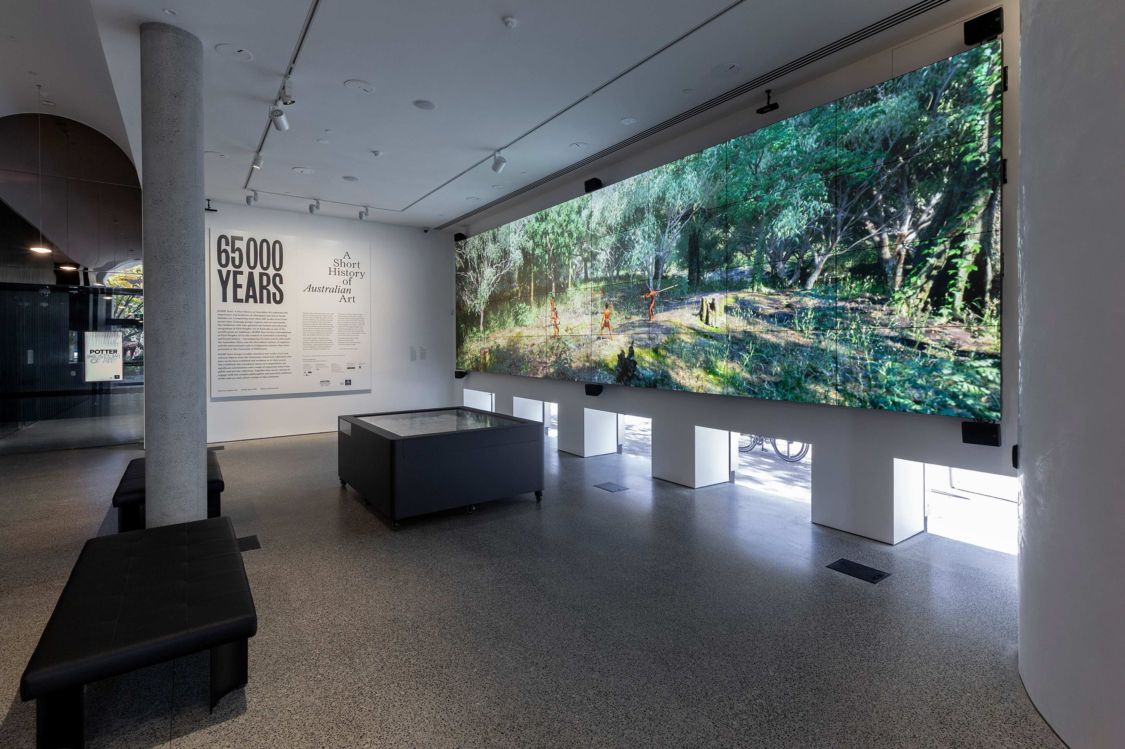 A bright gallery space features benches, a large wall display of lush green forest artwork, and a panel on the wall titled “65,000 Years” highlighting Aboriginal art and history.