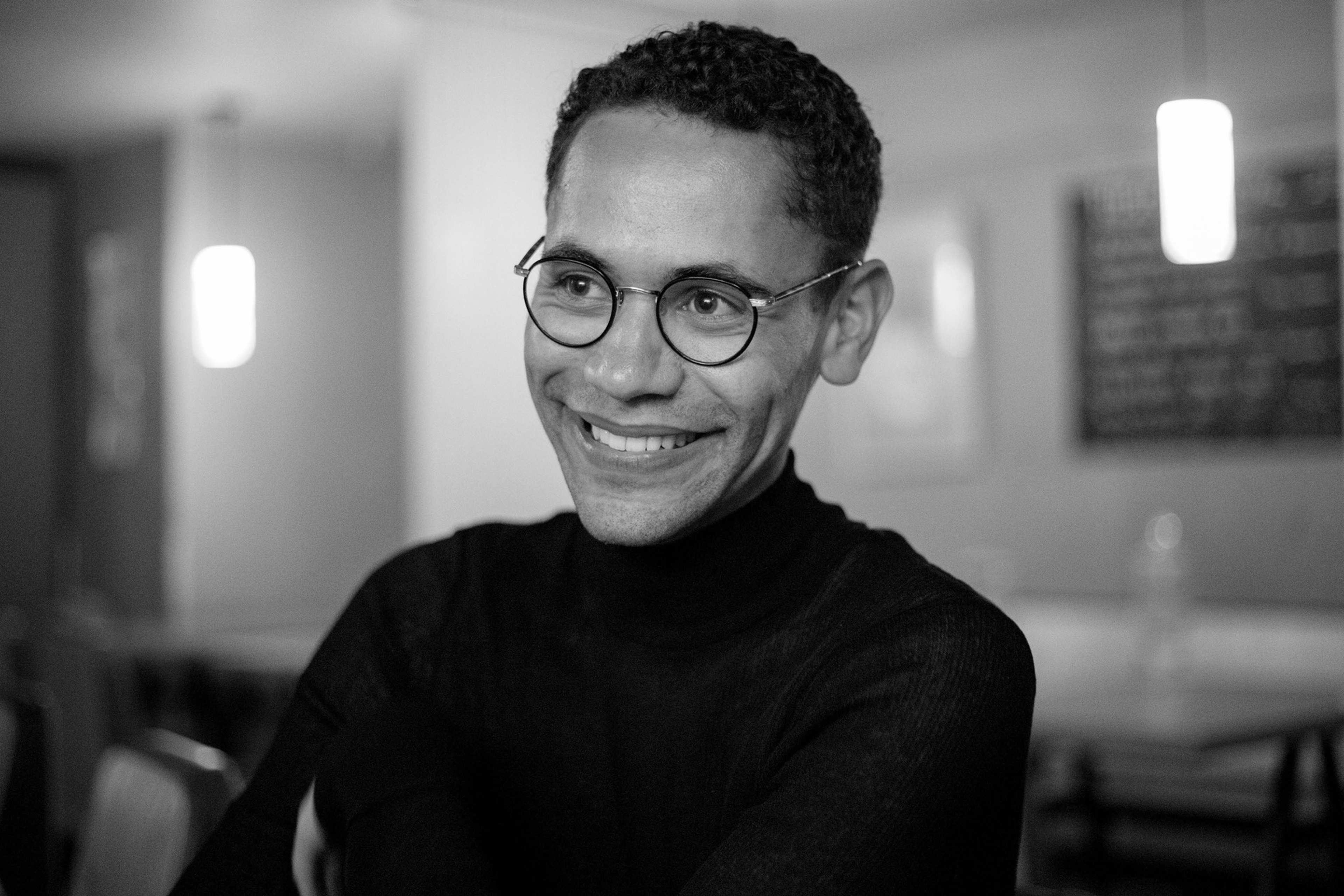 A person with short curly hair and glasses, wearing a dark turtleneck, smiles while sitting in a softly lit indoor space with blurred lights and furniture in the background.