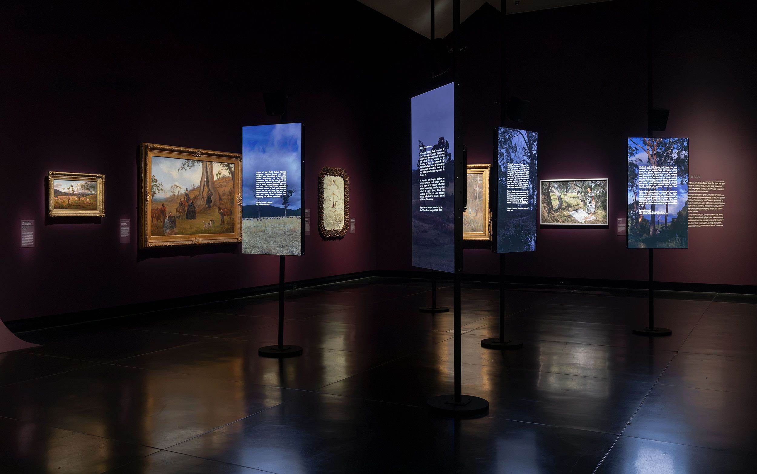 A dimly lit art gallery with framed paintings on dark purple walls. Four tall digital screens stand in the center, displaying text and images. The polished floor reflects the screens and artwork.