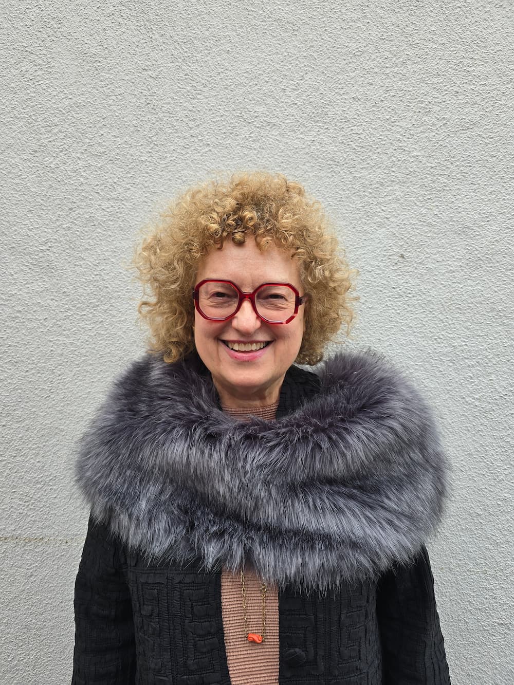 A smiling person with curly hair, red glasses, and a coat with a large fur collar stands beside a pink panel with the text “IN CONVERSATION WITH Carolyn Christov-Bakargiev.”.