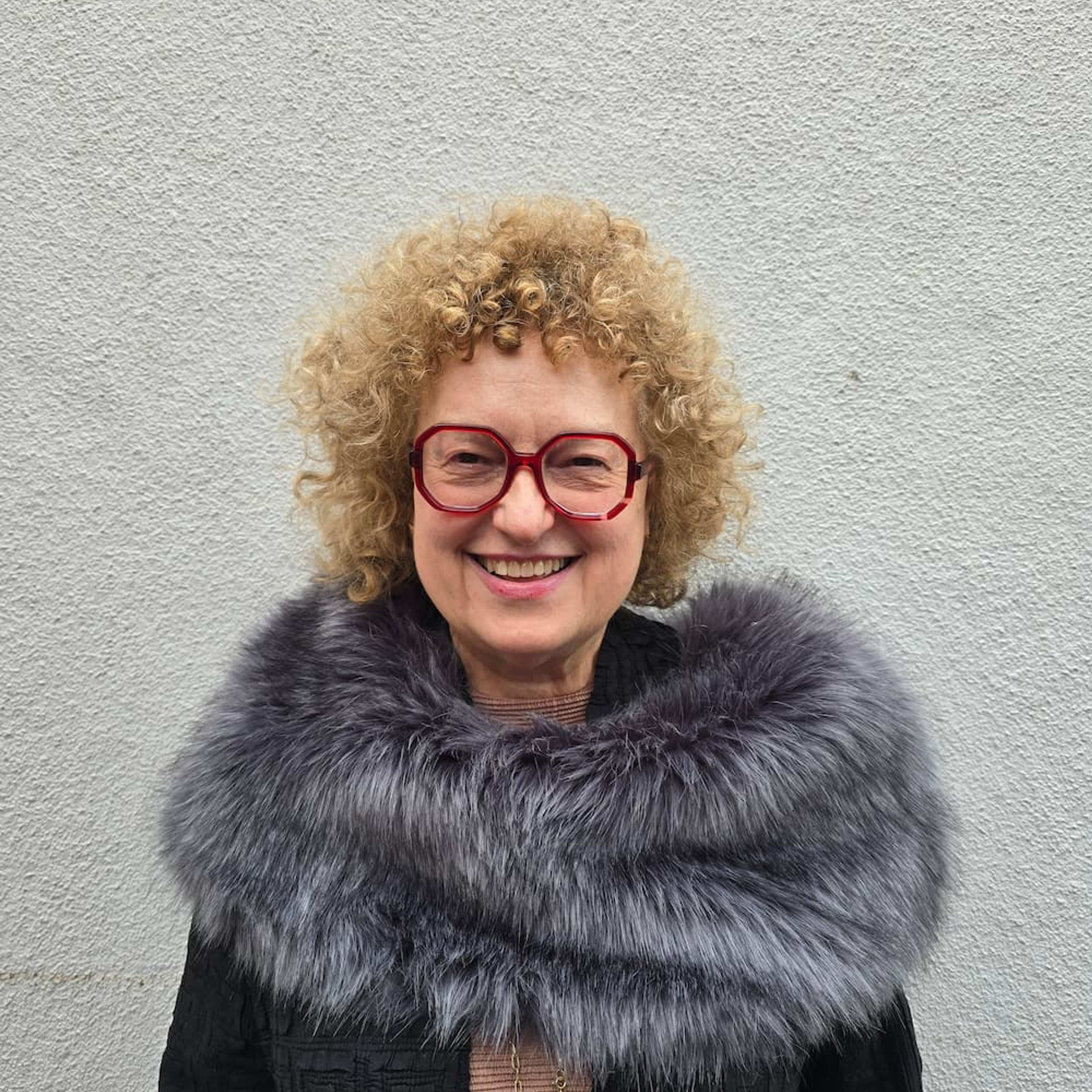 A smiling person with curly hair, red glasses, and a coat with a large fur collar stands beside a pink panel with the text “IN CONVERSATION WITH Carolyn Christov-Bakargiev.”.