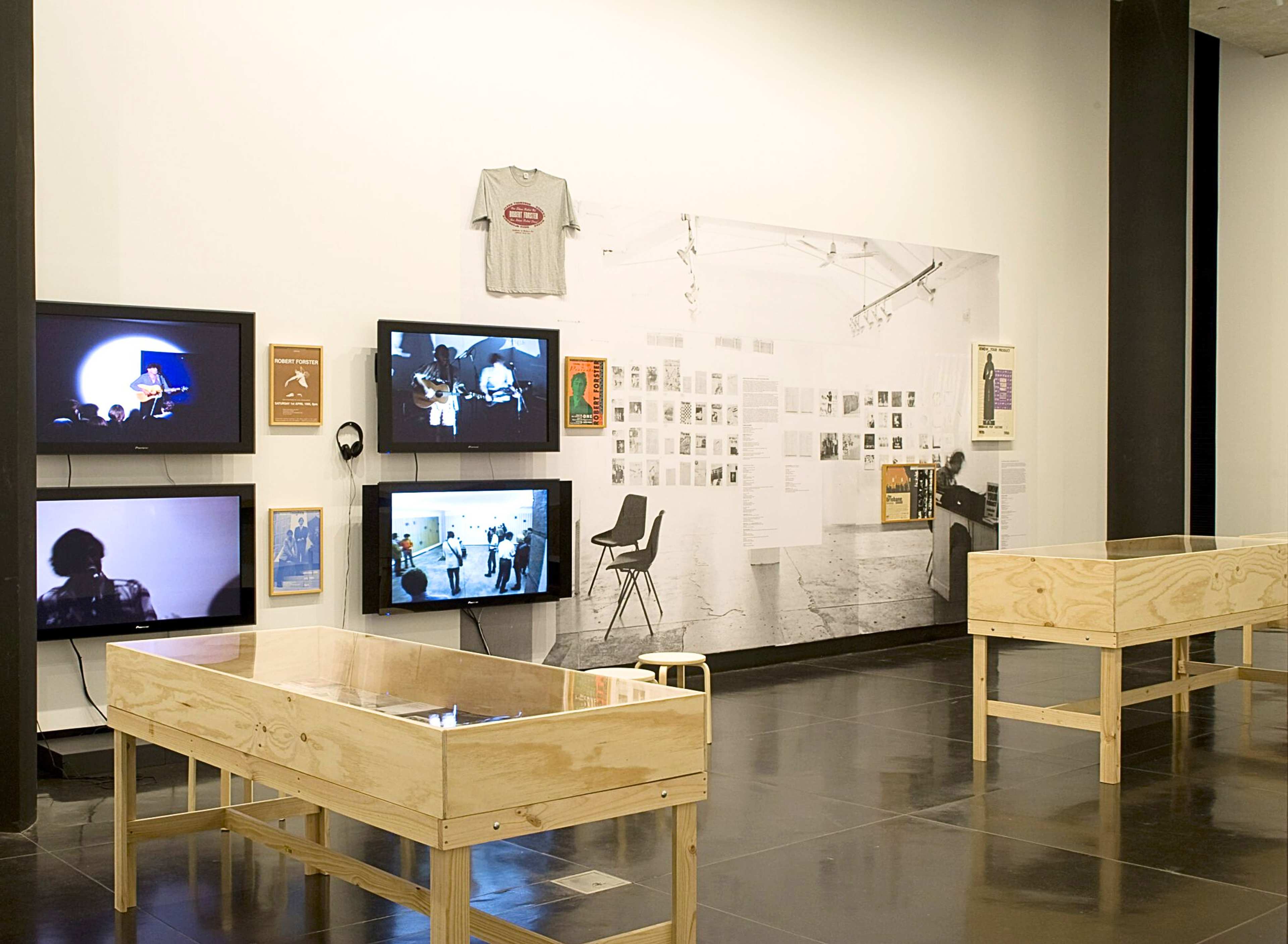 A photograph of a gallery with multiple vitrines, tv screens, a t-shirt and a large paste up on the wall