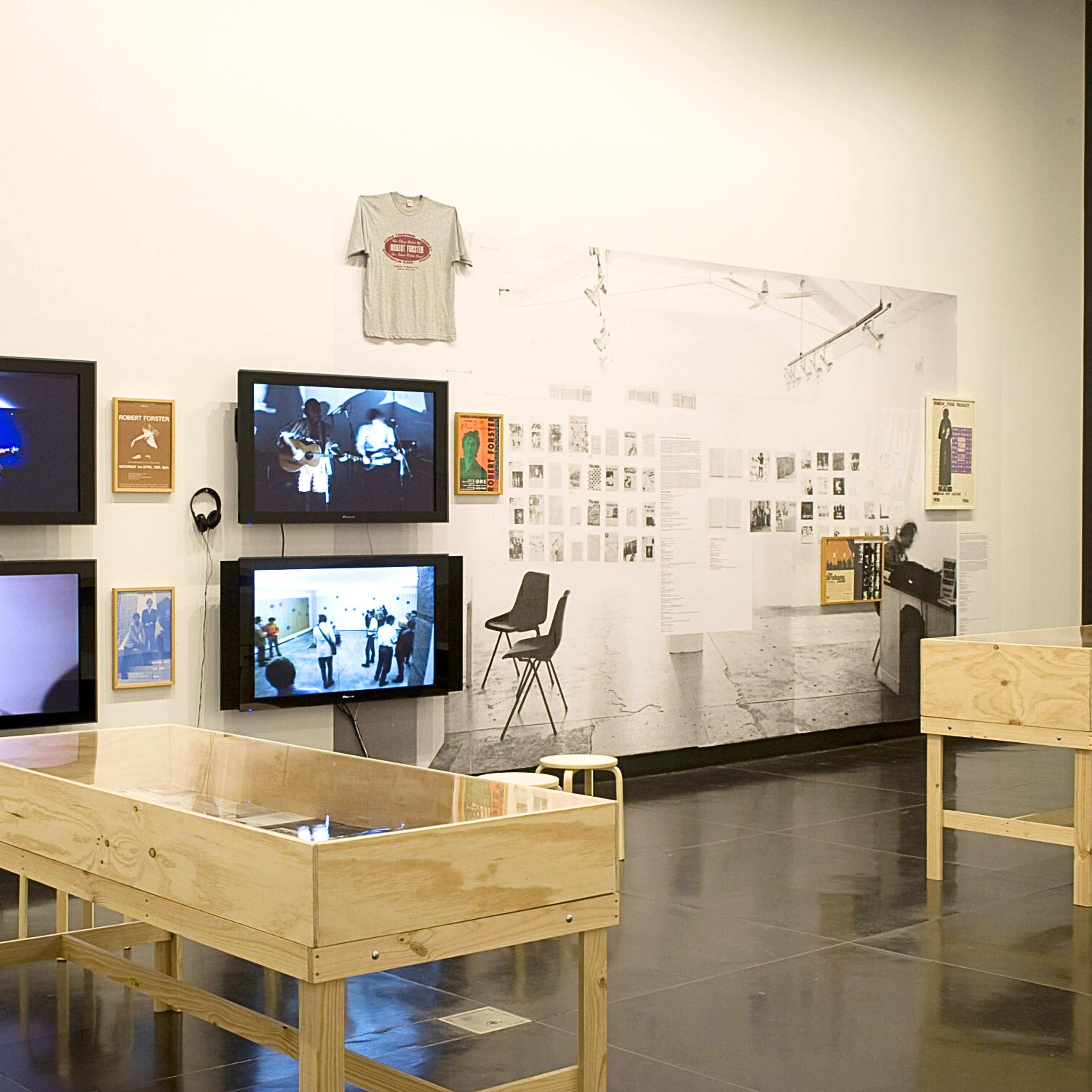 A photograph of a gallery with multiple vitrines, tv screens, a t-shirt and a large paste up on the wall