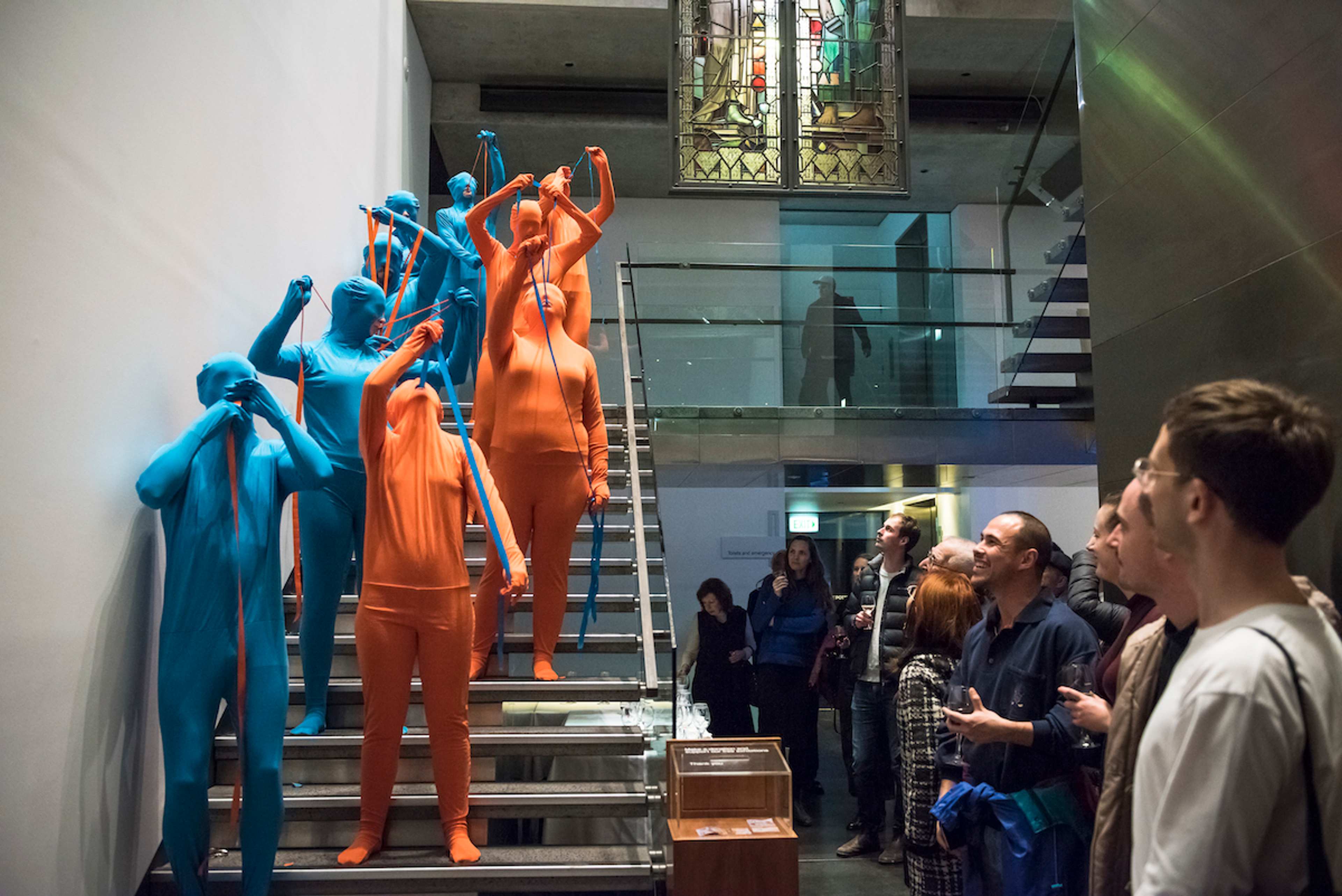 Performers in blue and orange body suits perform on a staircase as visitor watch