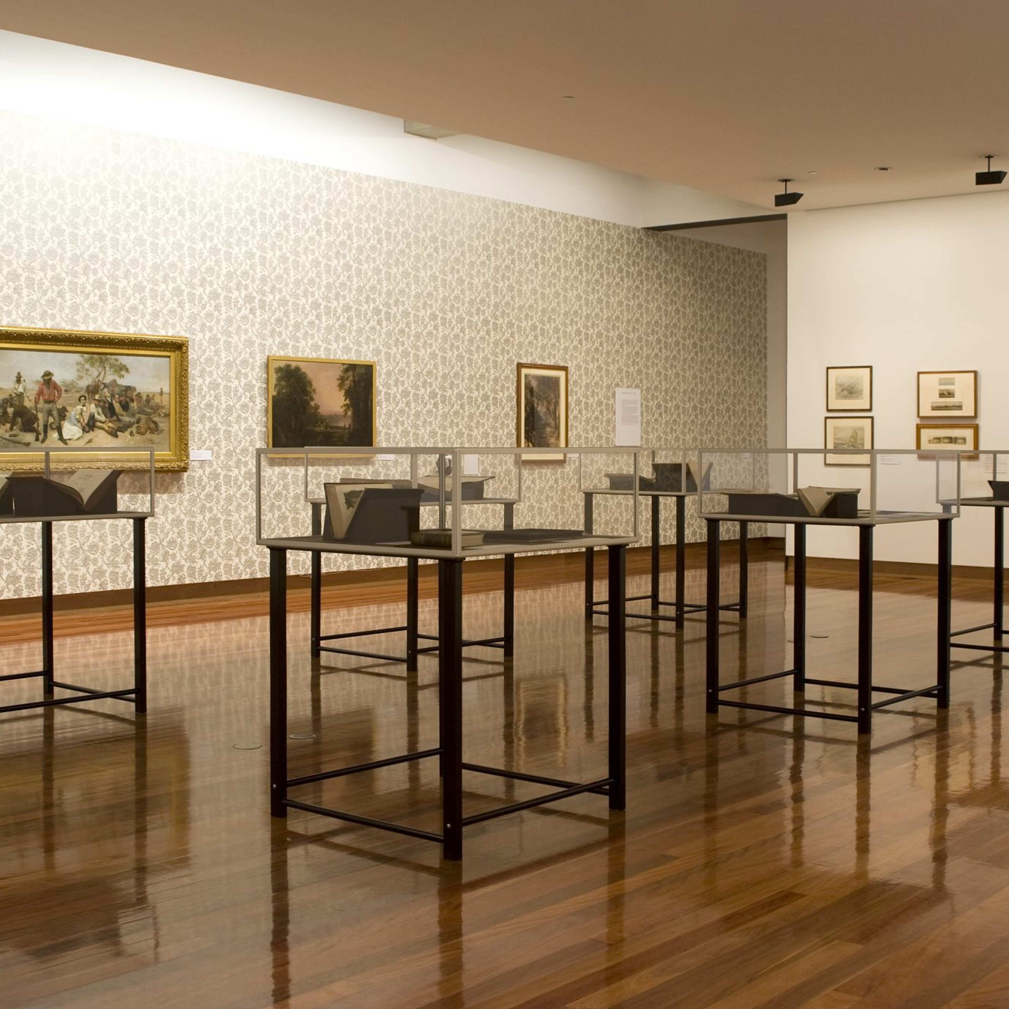 A photograph of a gallery space filled with smaller vitrines surrounded by paintings on the wallpapered walls
