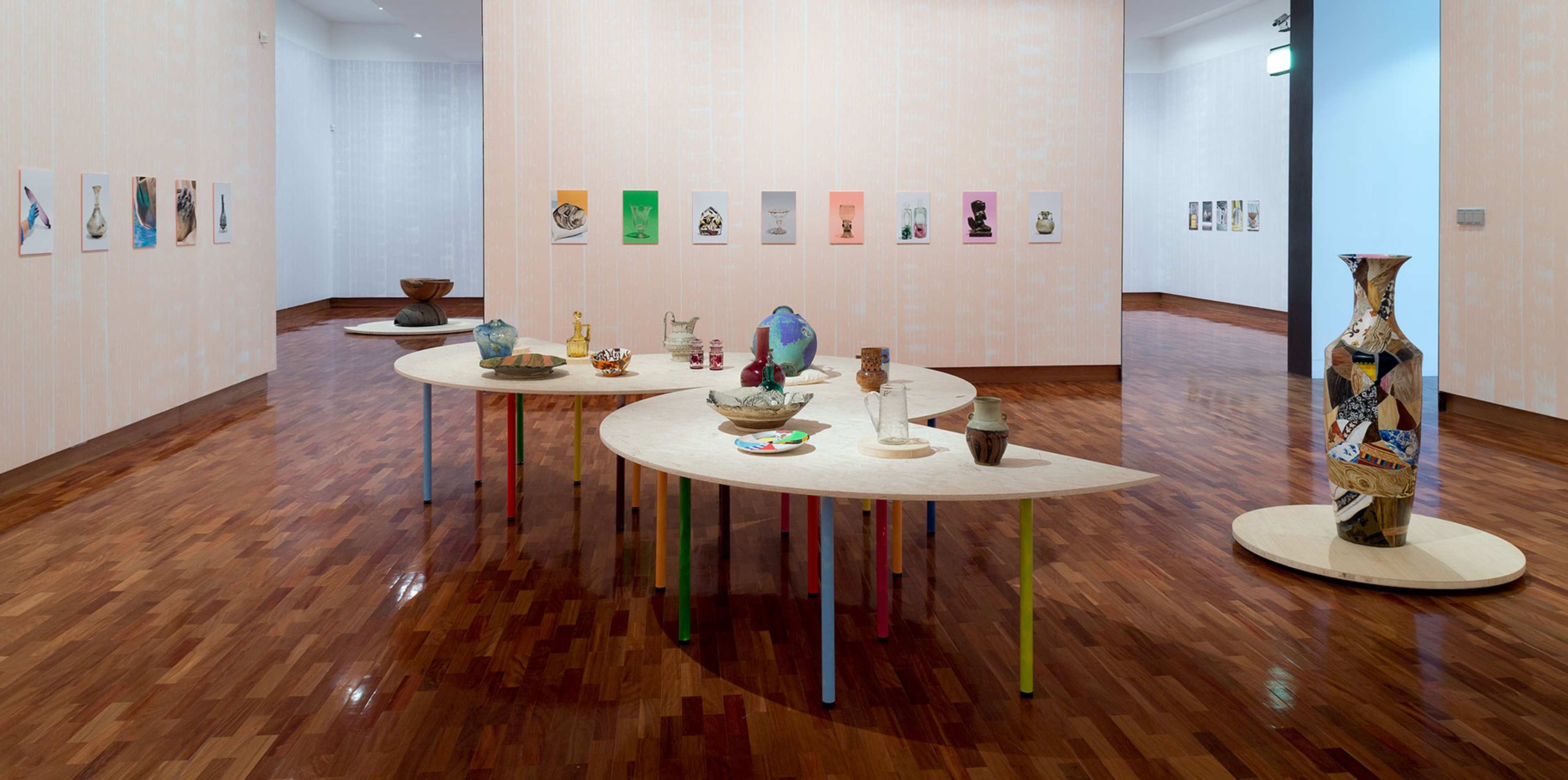 Plates bowls and decorative objects made from various materials are displayed on a plywood table with colourful prints hanging on the wall behind. A large human height colourful vase is positioned next to the tables on a low plinth,