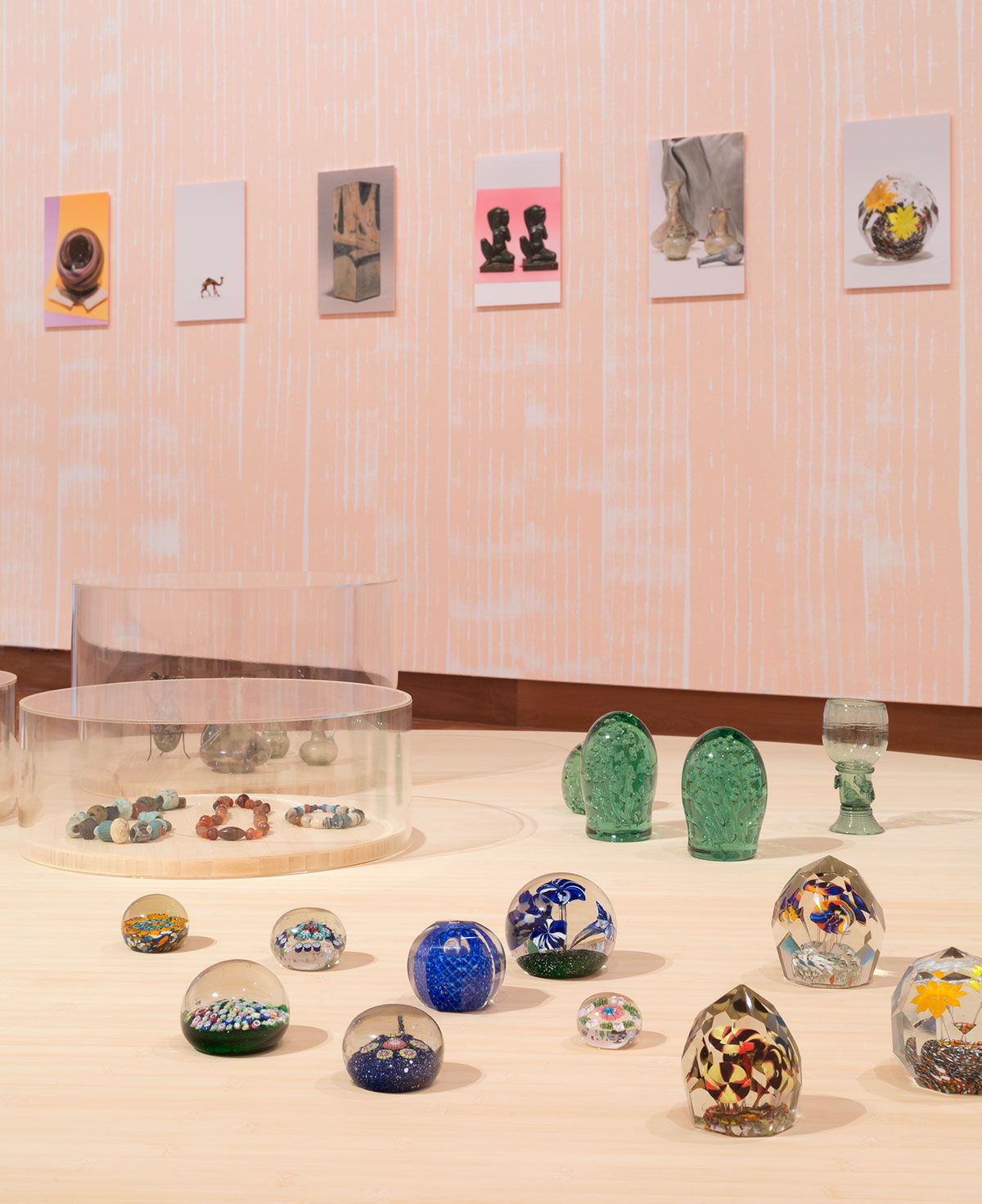 Snow domes and other glass decorative objects are displayed on plywood support with pink wall behind and several photographs of other objects in a row on that wall. 