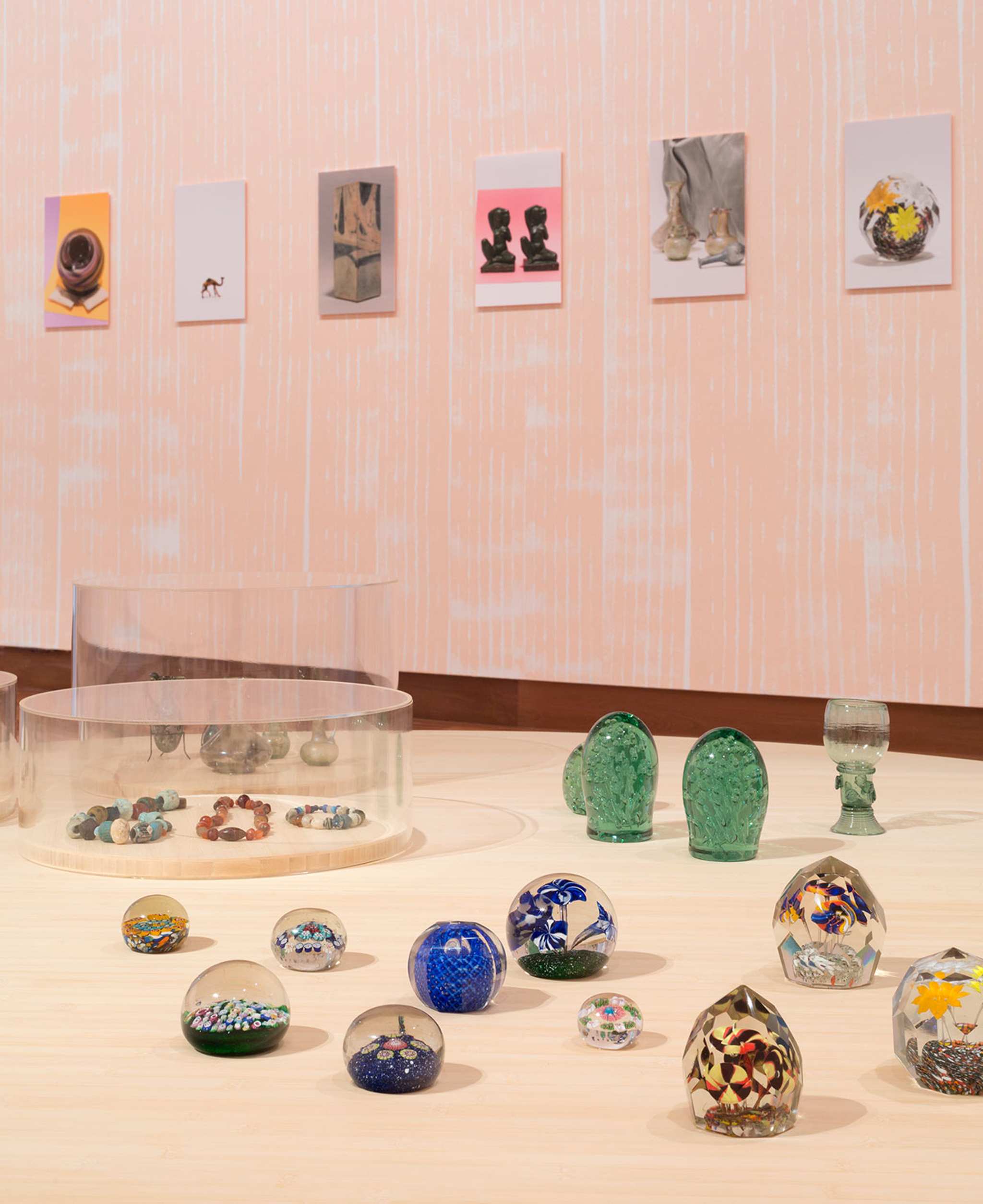 Snow domes and other glass decorative objects are displayed on plywood support with pink wall behind and several photographs of other objects in a row on that wall.