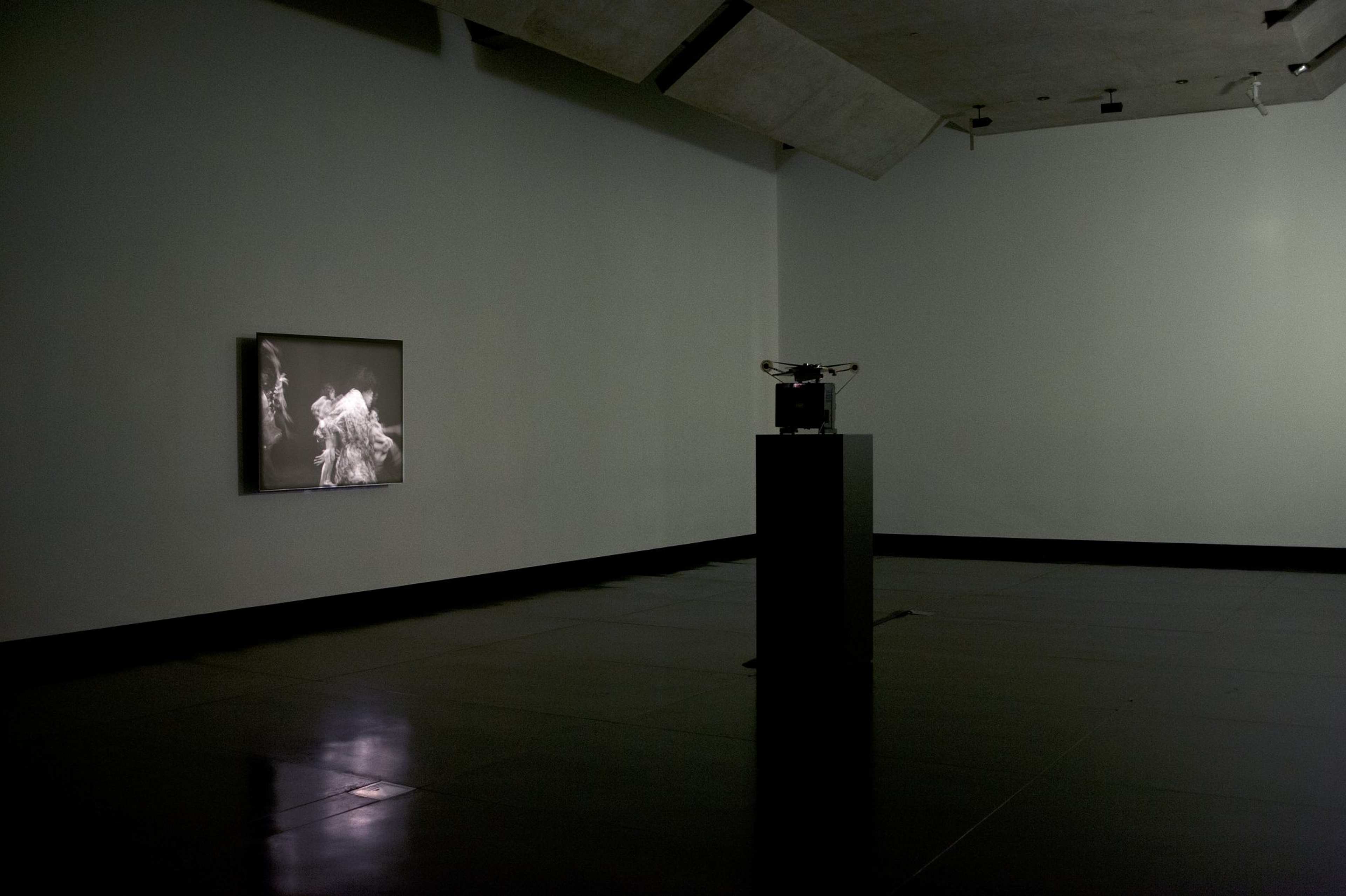 A photograph of a darkened gallery space with a 16 mm projector set on plinth in the middle, projecting a black and white image onto a wall-mounted screen.