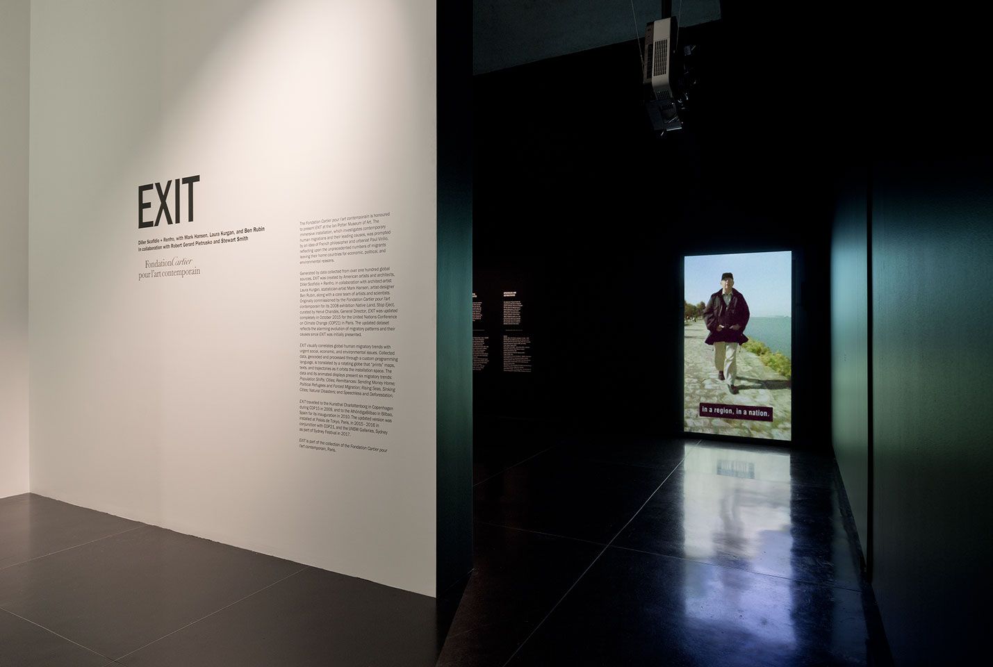 Entrance into a darkened gallery with exhibition title EXIT displayed on the wall to the left of the entry and a projection of a person walking towards the visitor on the wall ahead