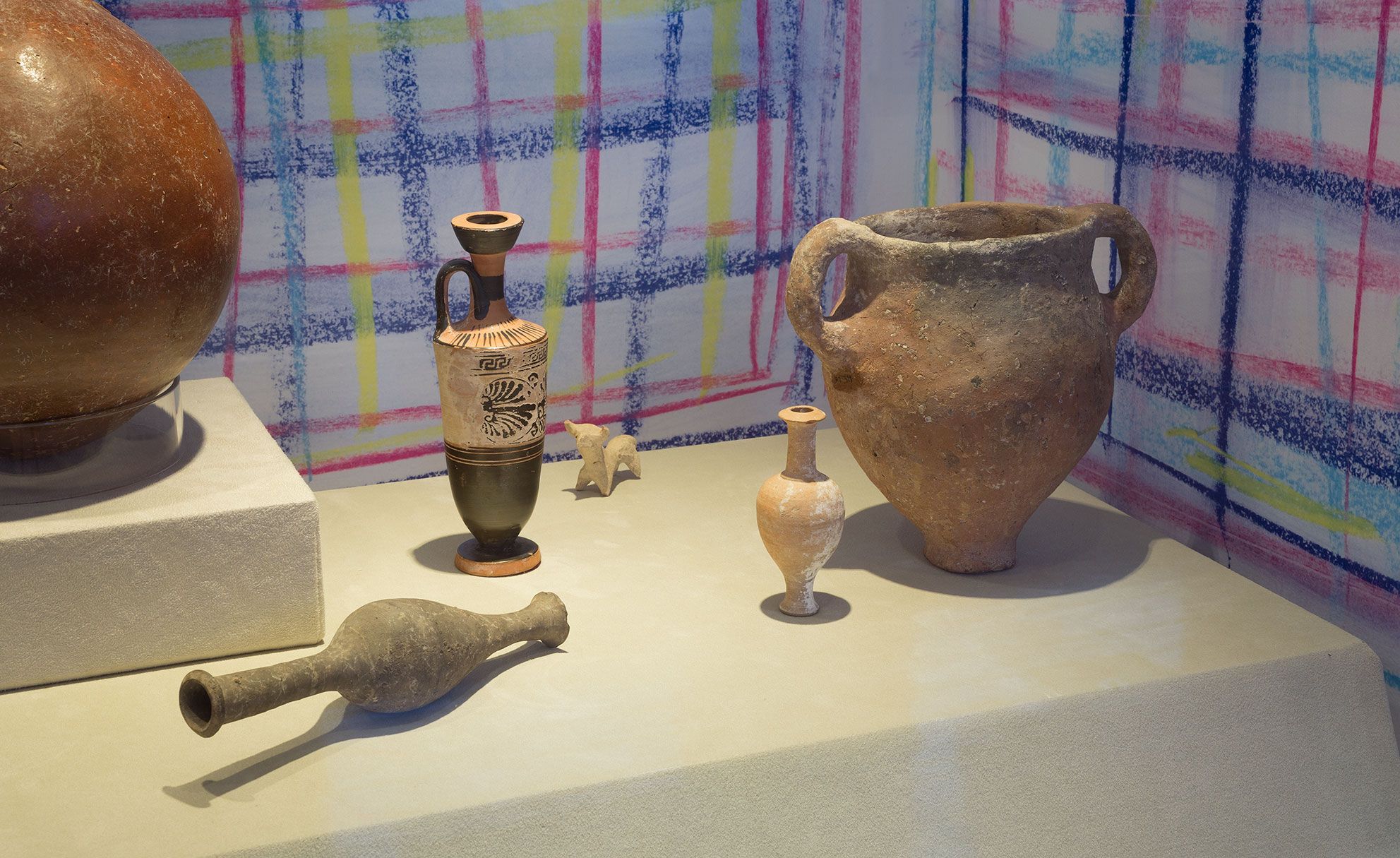 A photograph of ceramic ancient vessels of varying sizes arranged on a plinths against a colourful drawn background of blue, yellow and red lines 