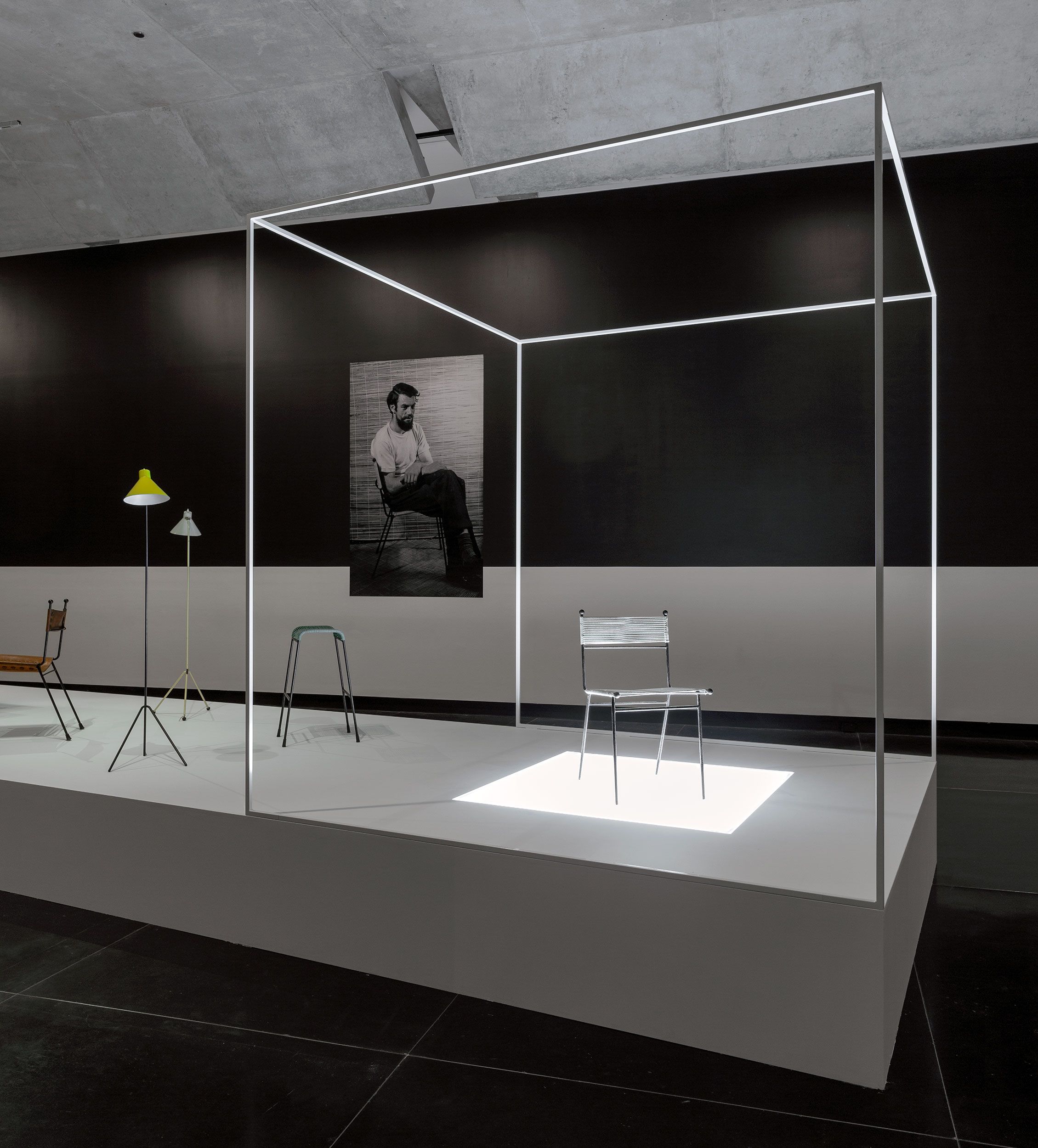 A photograph of a gallery space with mid-century furniture on a large plinth in the foreground and a black and white photograph of a seated person on the wall behind