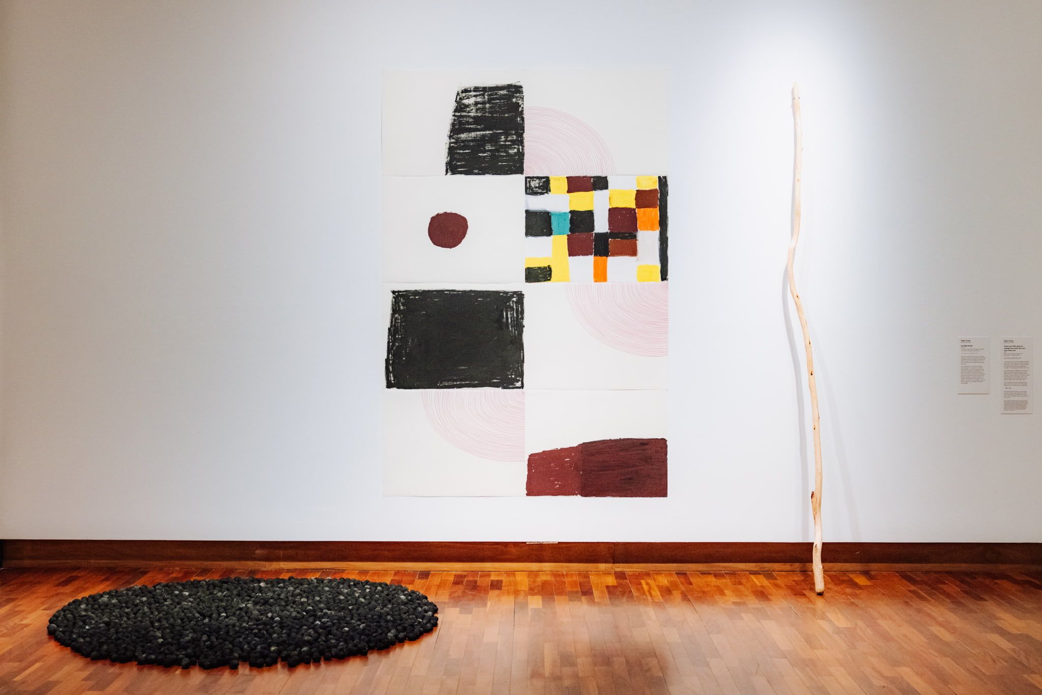 A white gallery wall displays abstract art: a large canvas with geometric shapes and colors, a tall, thin wooden stick leaning against the wall, and scattered small black objects on the wooden floor below.