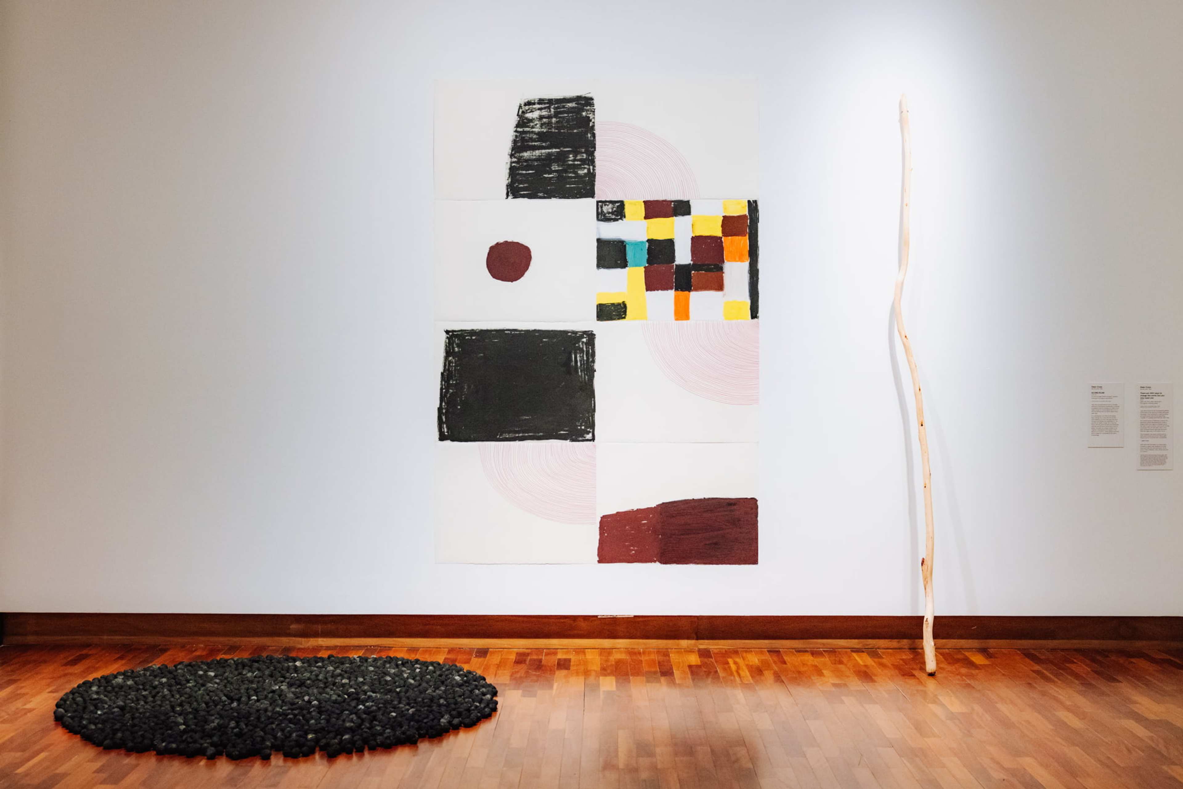 A white gallery wall displays abstract art: a large canvas with geometric shapes and colors, a tall, thin wooden stick leaning against the wall, and scattered small black objects on the wooden floor below.