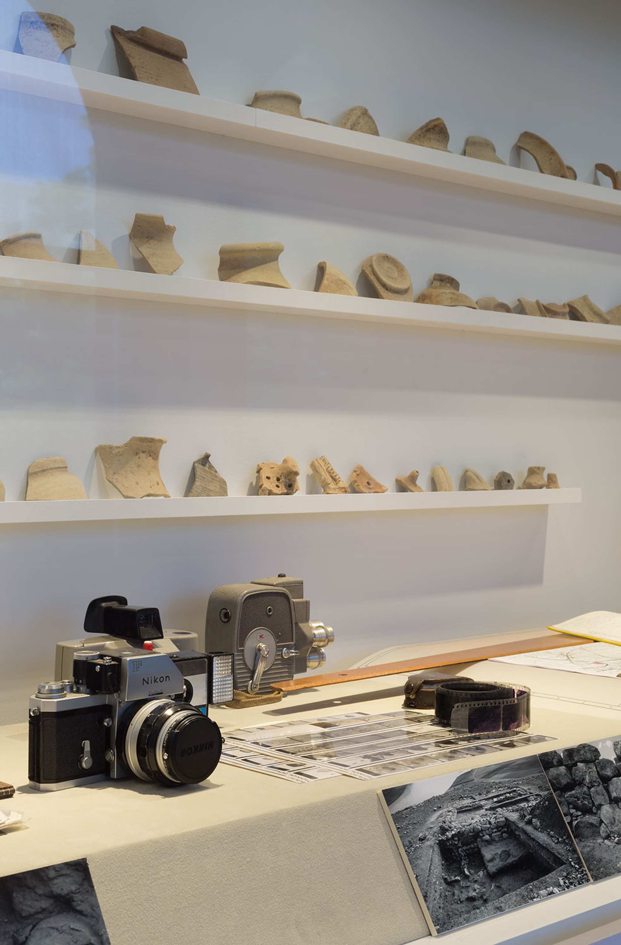 A photograph of a gallery with a large vitrine displaying a large array of ancient ceramics and a film photographic camera and some black and white photographs in the foreground