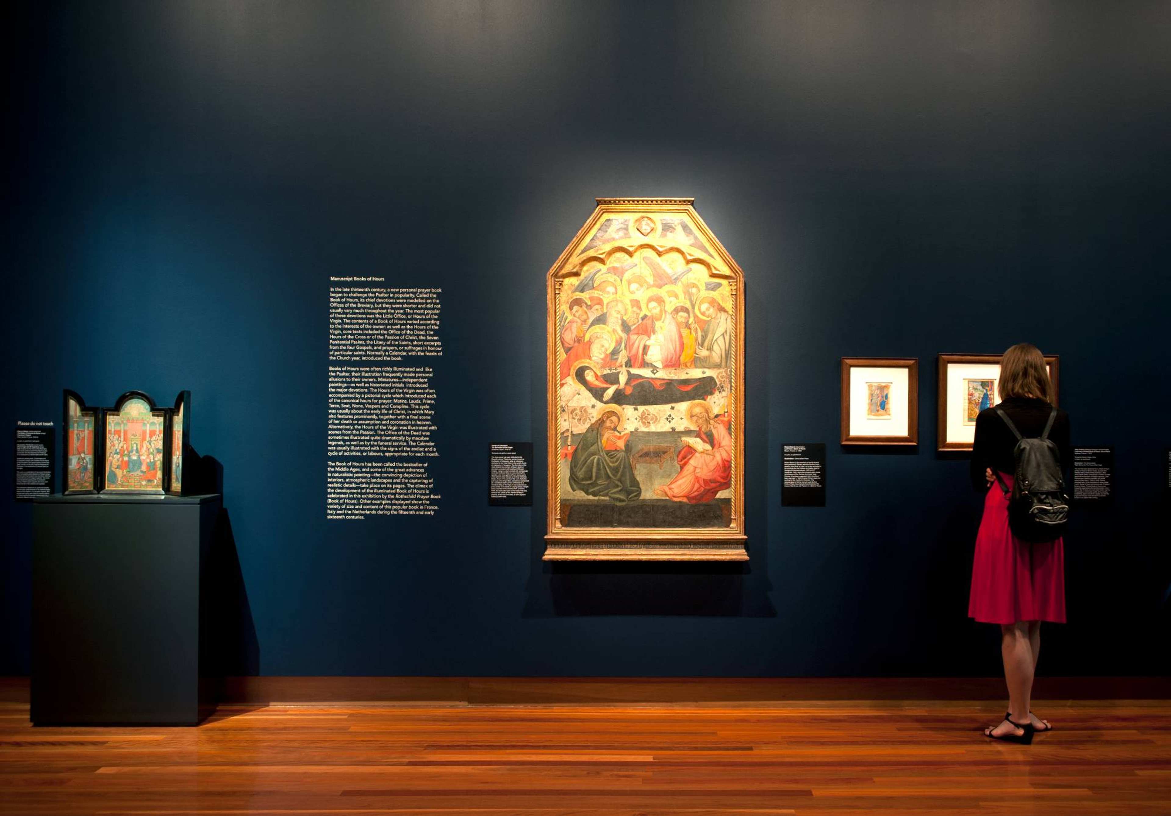 A dark gallery with  large medieval painting on a black-painted wall surrounded by smaller works