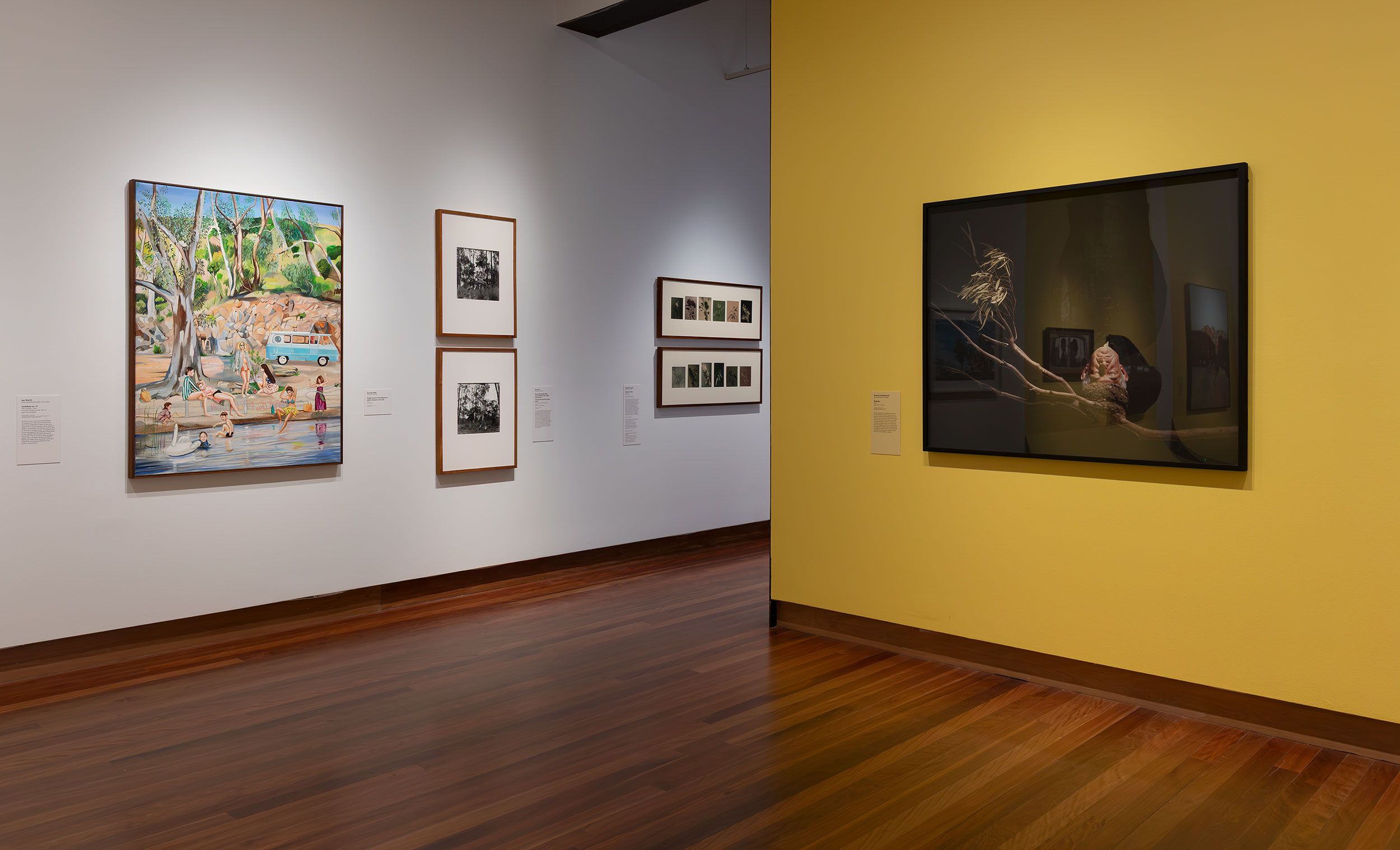An art gallery interior with wooden floors, white and yellow walls, displaying colorful paintings and black-and-white photographs in frames. The space is well-lit and organized, creating a calm and inviting atmosphere.
