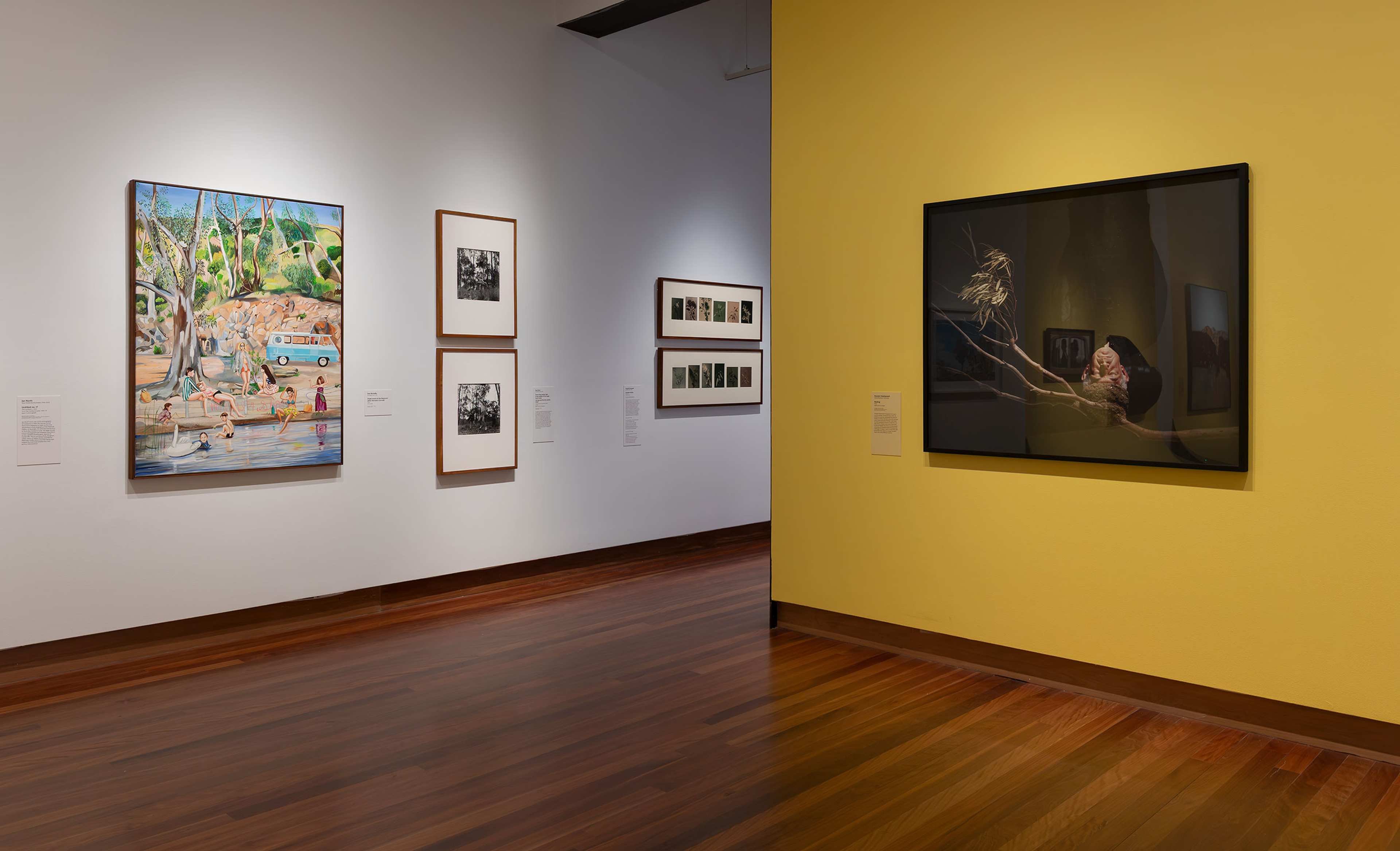 An art gallery interior with wooden floors, white and yellow walls, displaying colorful paintings and black-and-white photographs in frames. The space is well-lit and organized, creating a calm and inviting atmosphere.