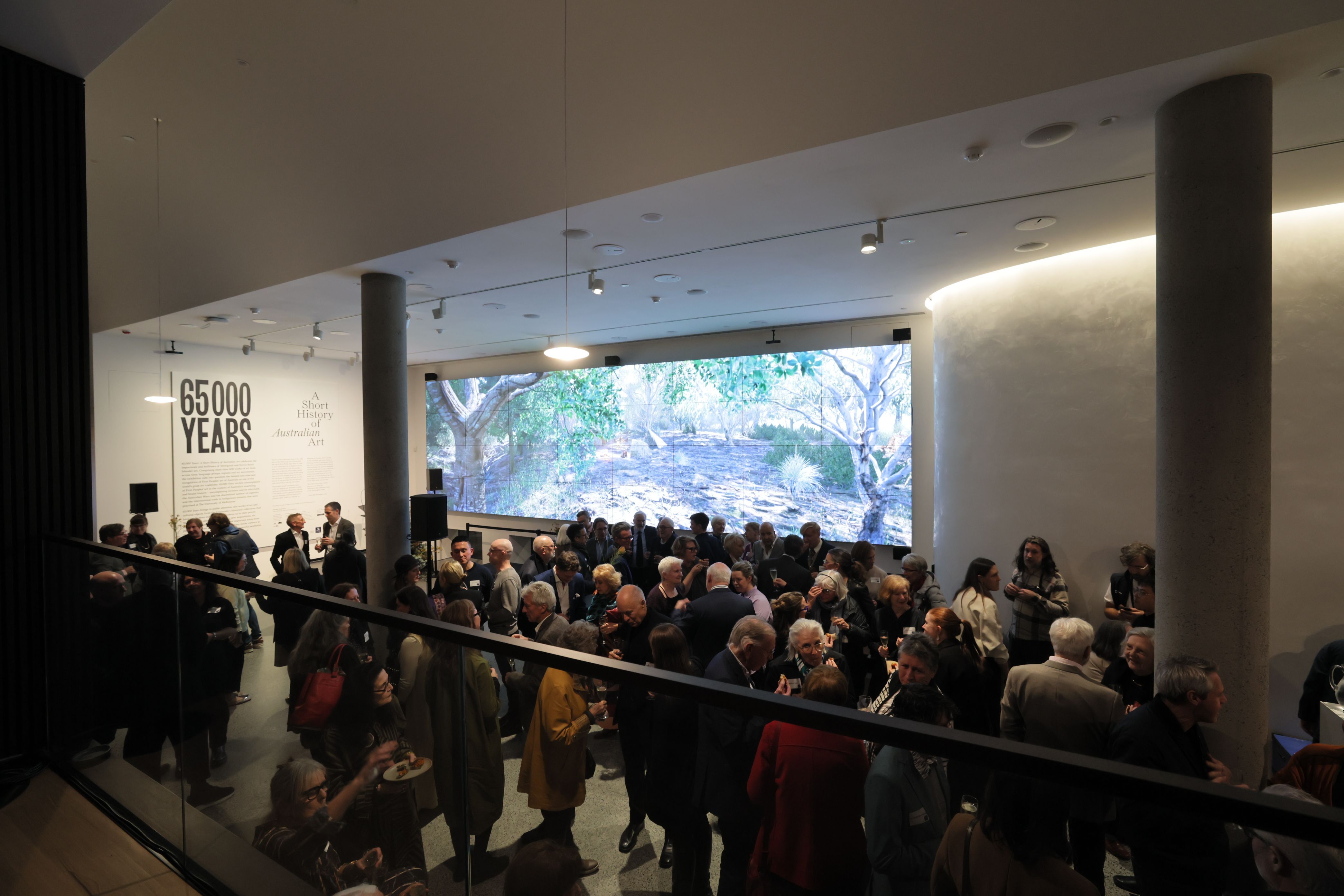 A large group of people gather in a modern indoor space with floor-to-ceiling artwork showing a forest scene. The wall features the text 65,000 YEARS. The atmosphere is lively and social.