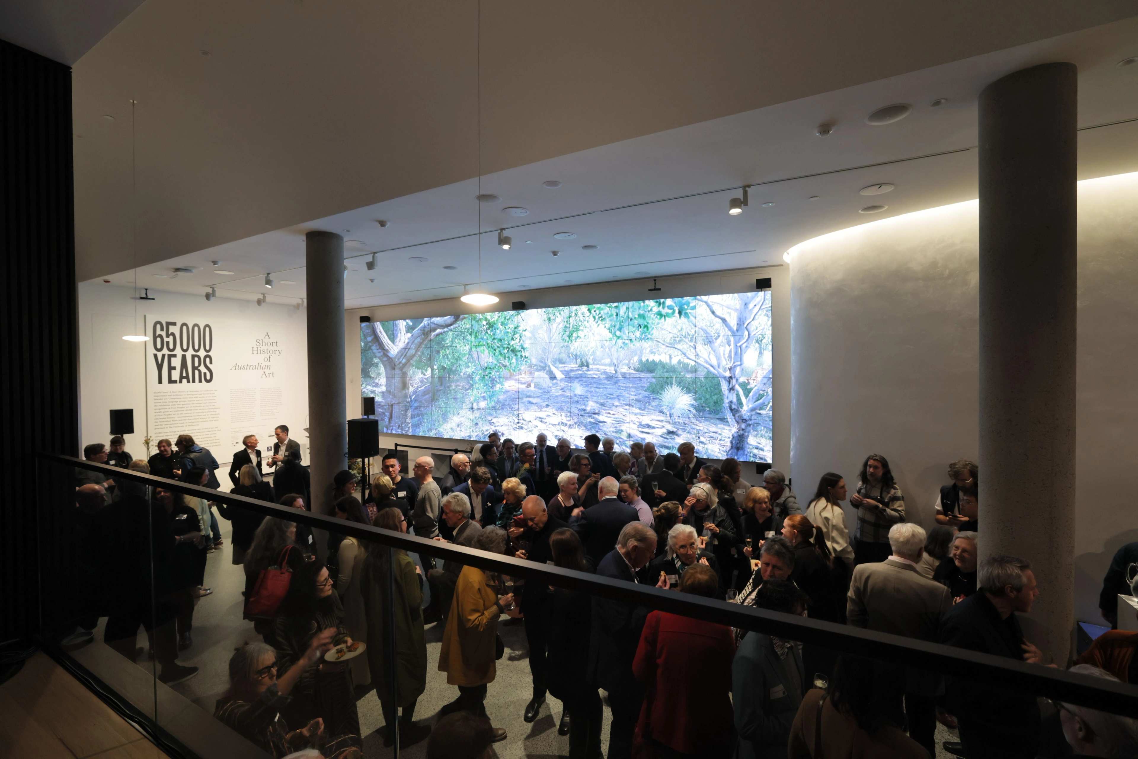 A large group of people gather in a modern indoor space with floor-to-ceiling artwork showing a forest scene. The wall features the text 65,000 YEARS. The atmosphere is lively and social.