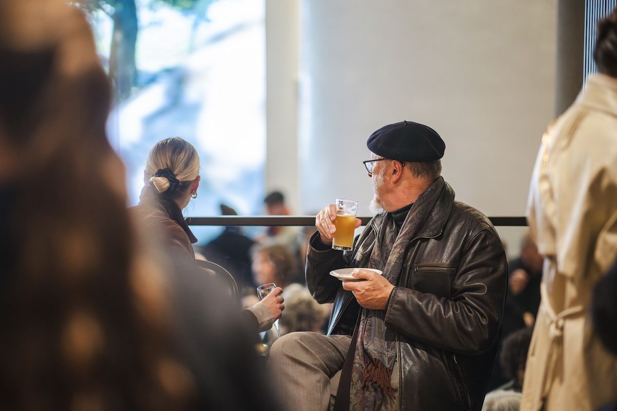 An older man in a beret and leather jacket drinks from a glass while sitting indoors. He faces a woman with blond hair in a bun. The setting is busy, with blurred people in the background.