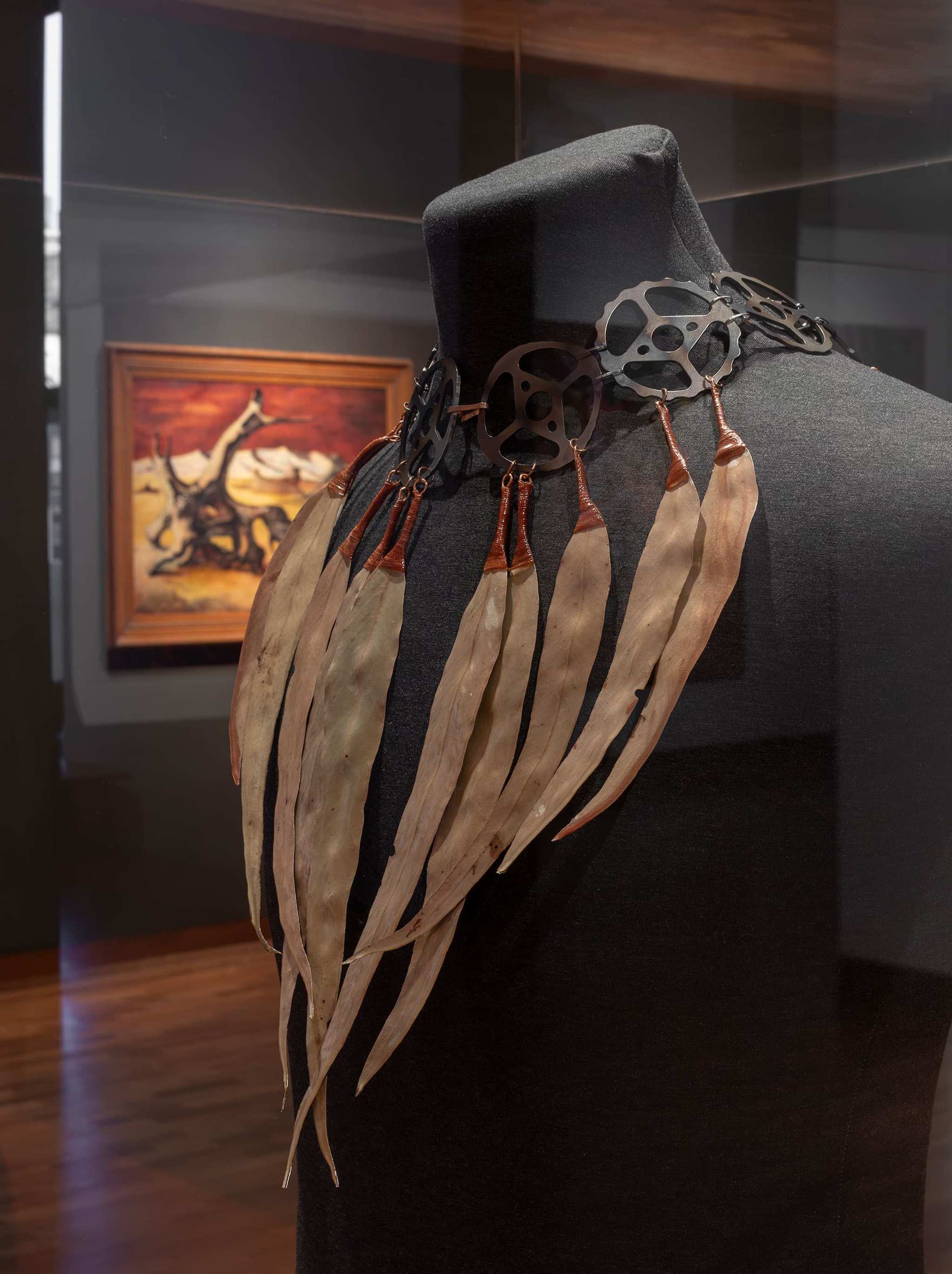 A mannequin displays a necklace made of black metal gears and long, curved, leaf-like elements. In the background, a framed painting with warm tones hangs on a dark wall.