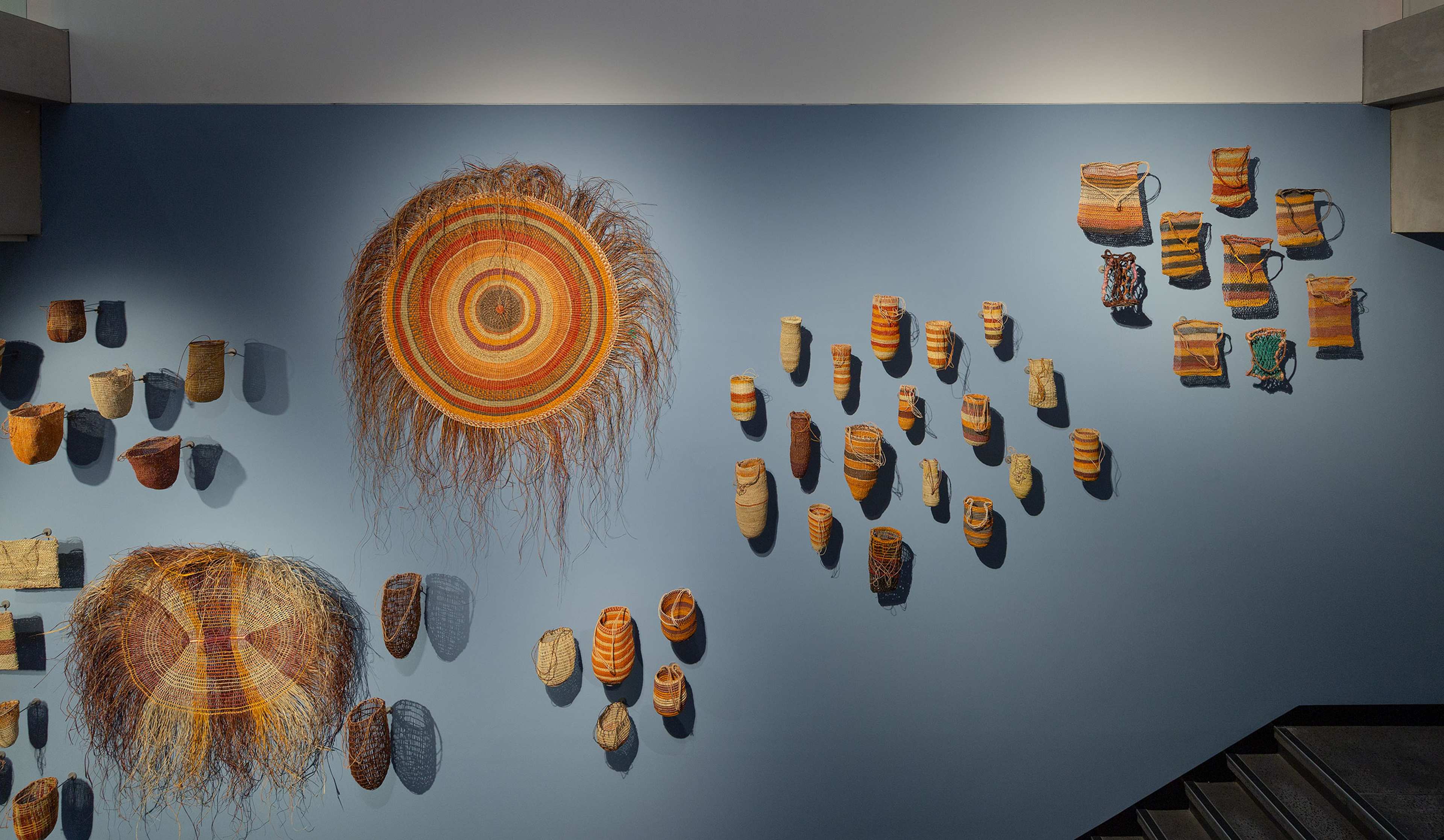 Woven baskets and large, circular woven art pieces are displayed on a blue wall, arranged in clusters and rows, with stairs visible at the bottom right corner.