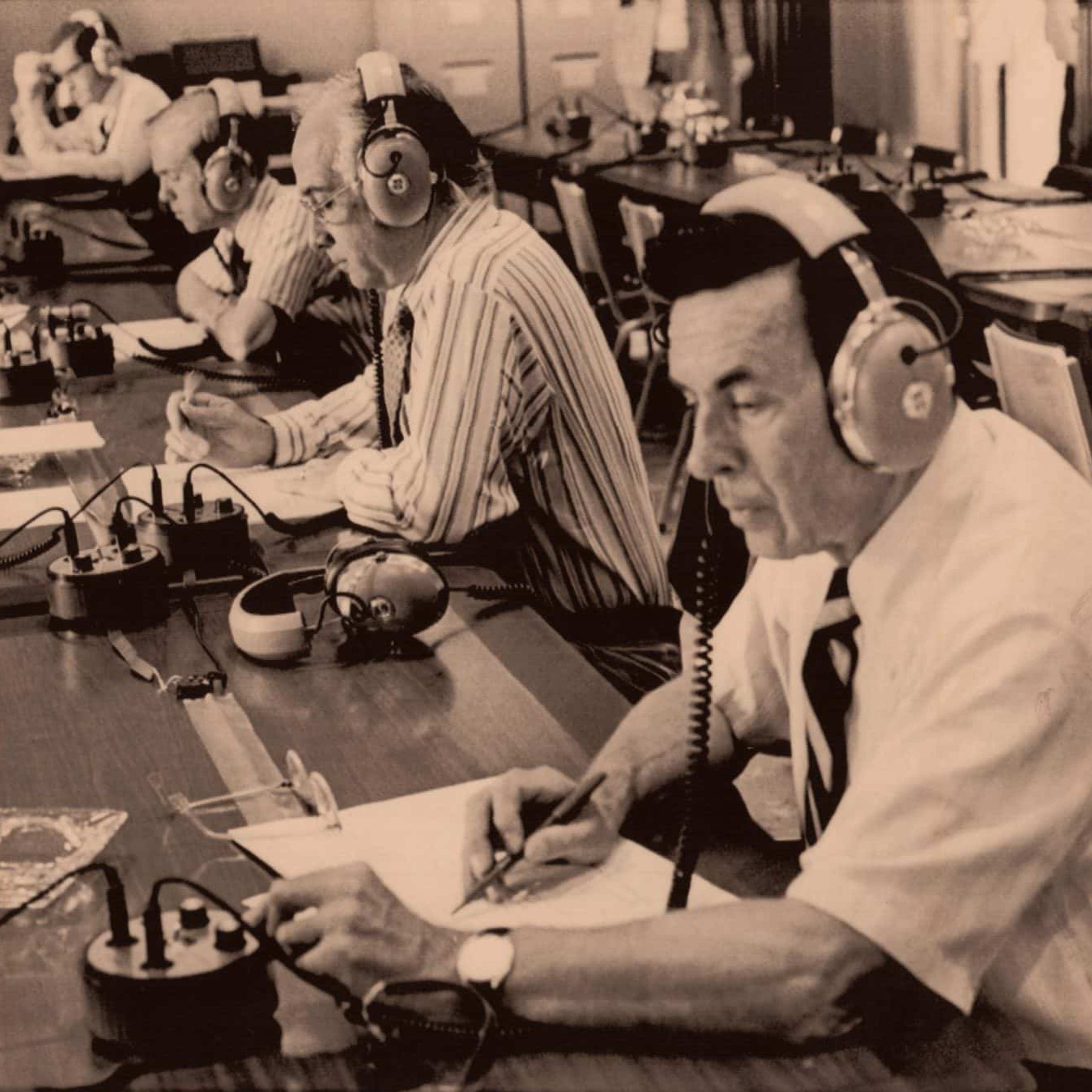A black and white photo of a row of men with headphones on writing notes on a piece of paper