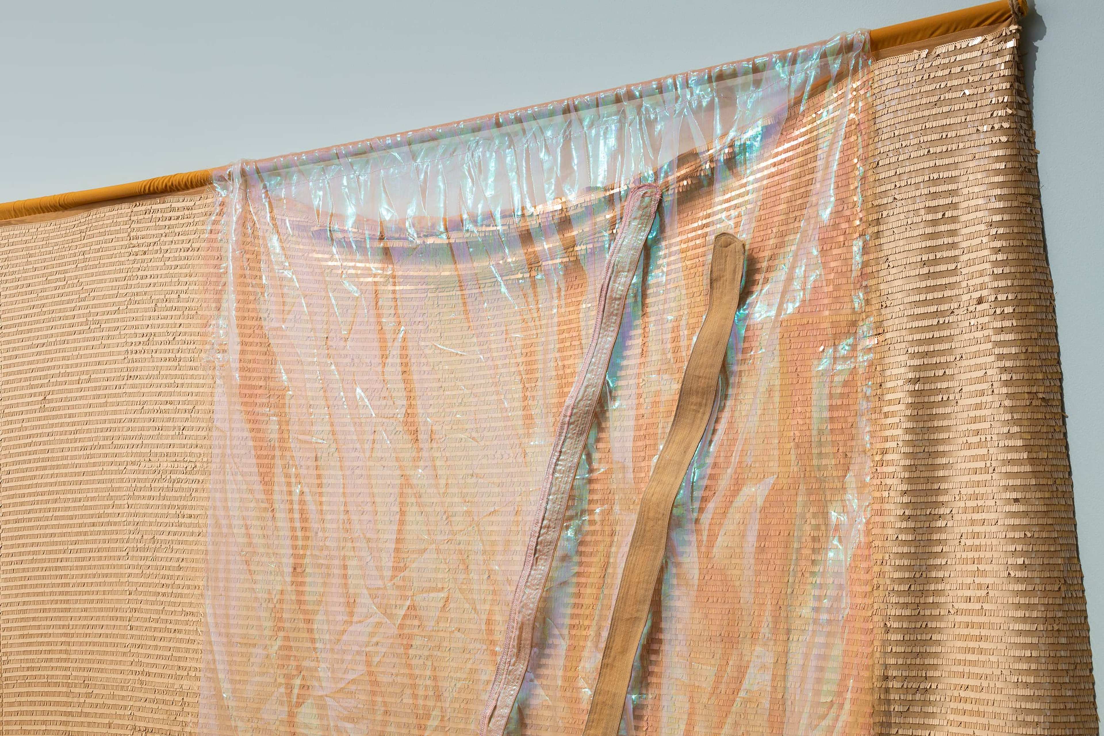 A beige woven curtain with a sheer, iridescent plastic sheet draped over it, attached to a yellow rod against a light gray wall. A beige fabric strap hangs loosely on the right side.